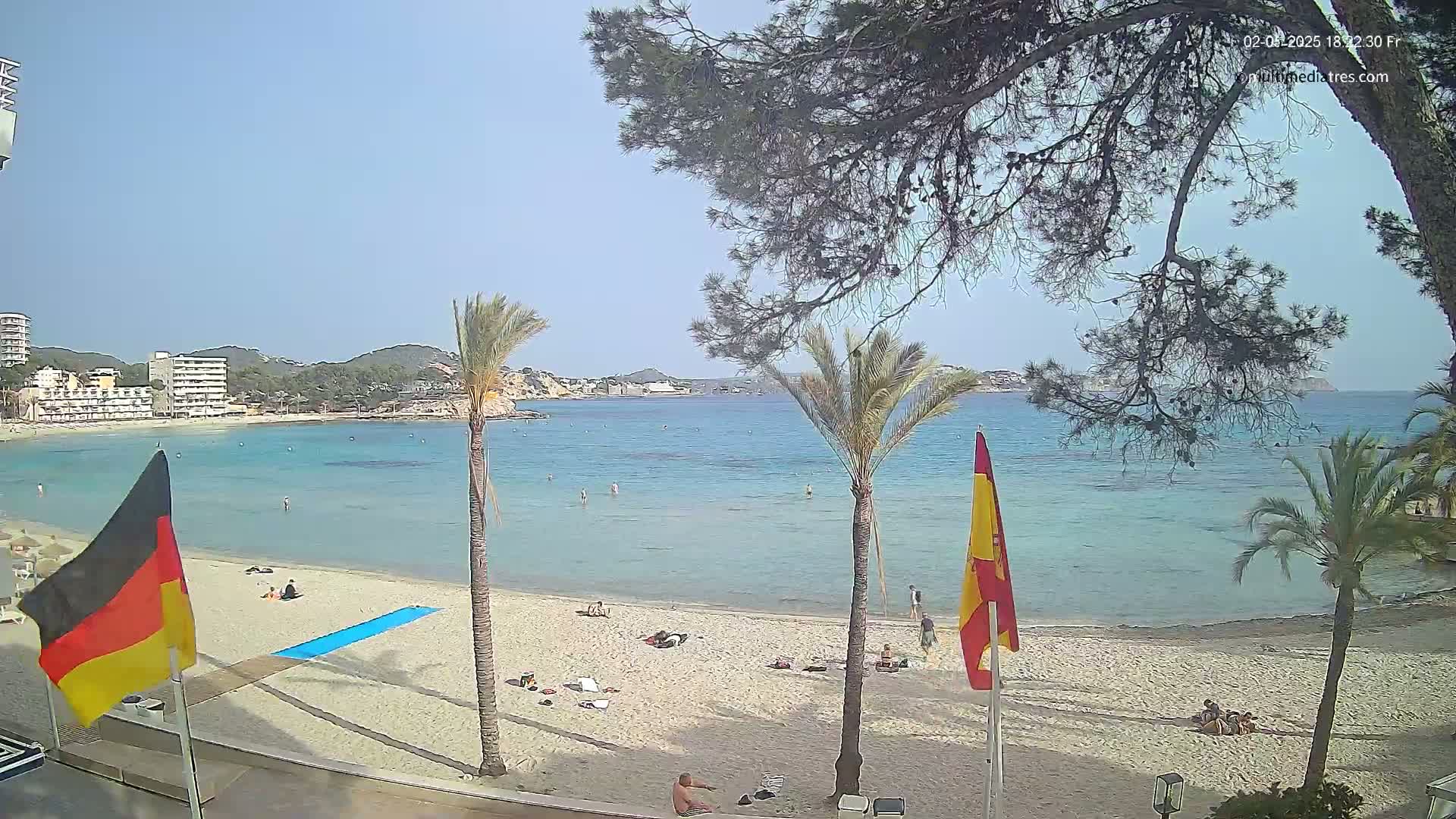A sandy beach with clear, shallow turquoise water, palm trees, and several people relaxing, under a sunny sky.