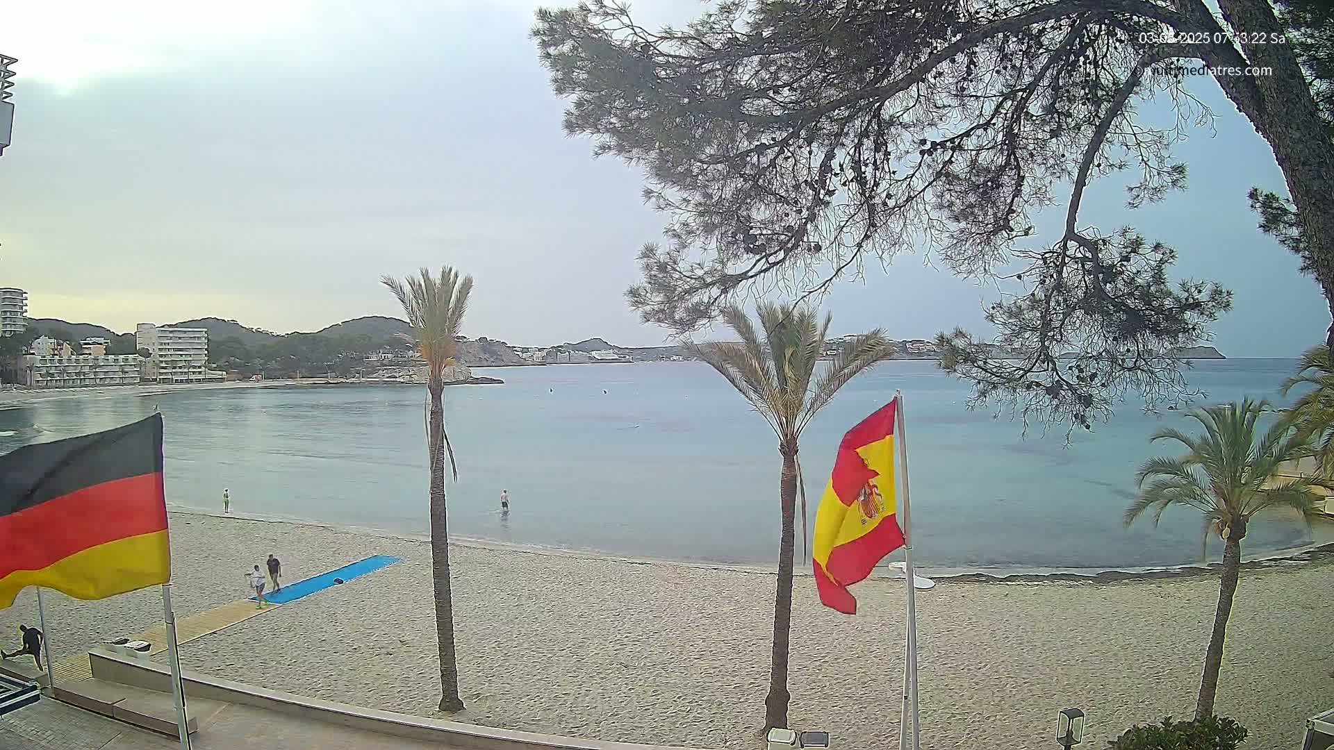 A sandy beach with calm, light blue water, palm trees, and flags of Germany and Spain is visible under an overcast sky.