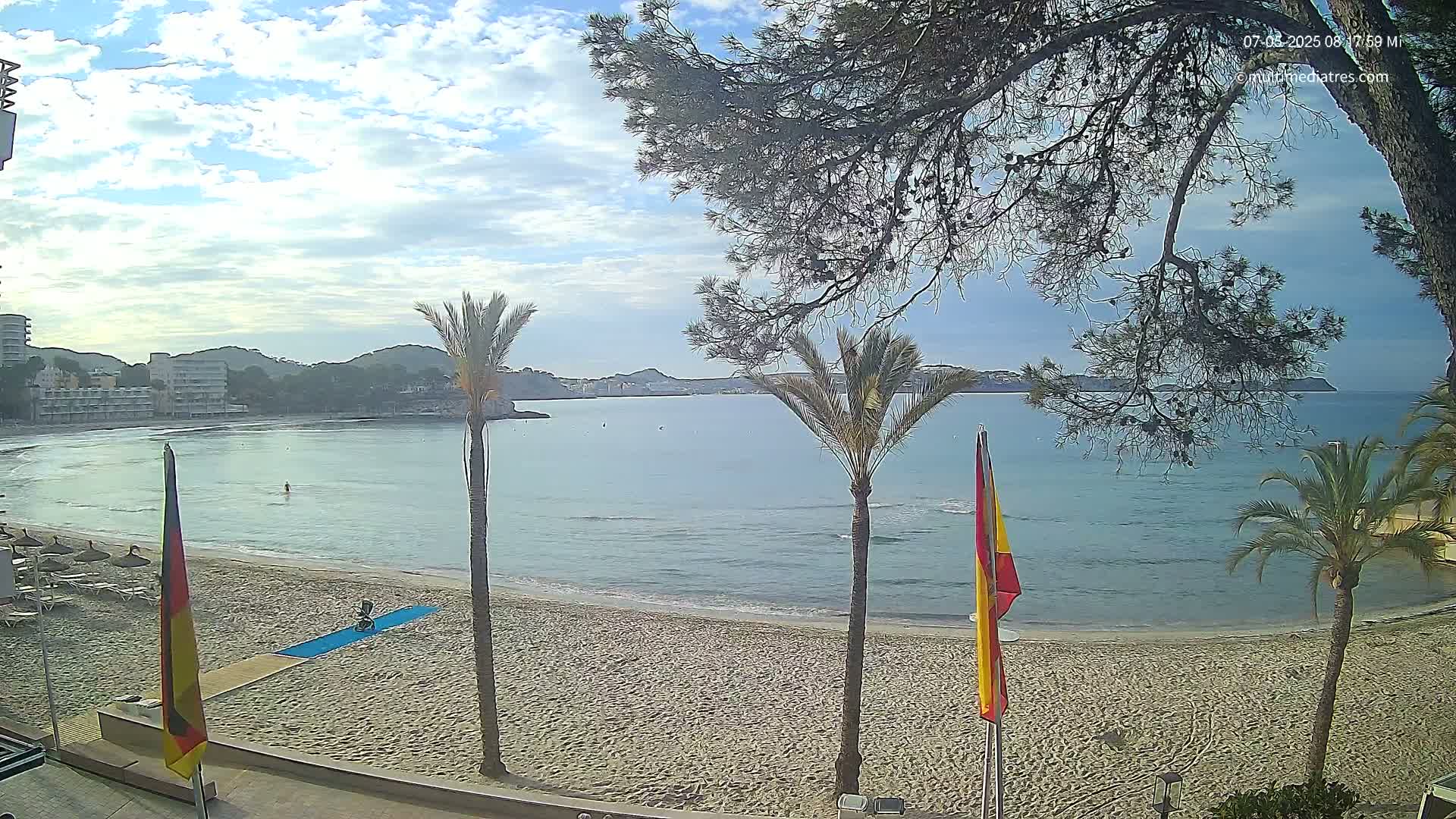 A partly cloudy day shows a sandy beach with calm, light-blue water, palm trees, and buildings in the background.
