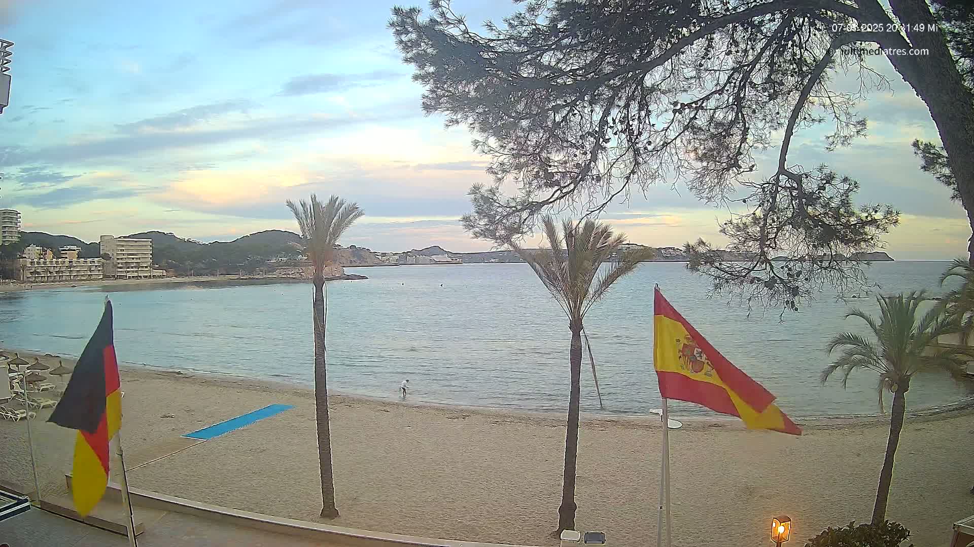 A calm, pale-blue ocean bay with a sandy beach, palm trees, and buildings in the background under a partly cloudy, twilight sky.