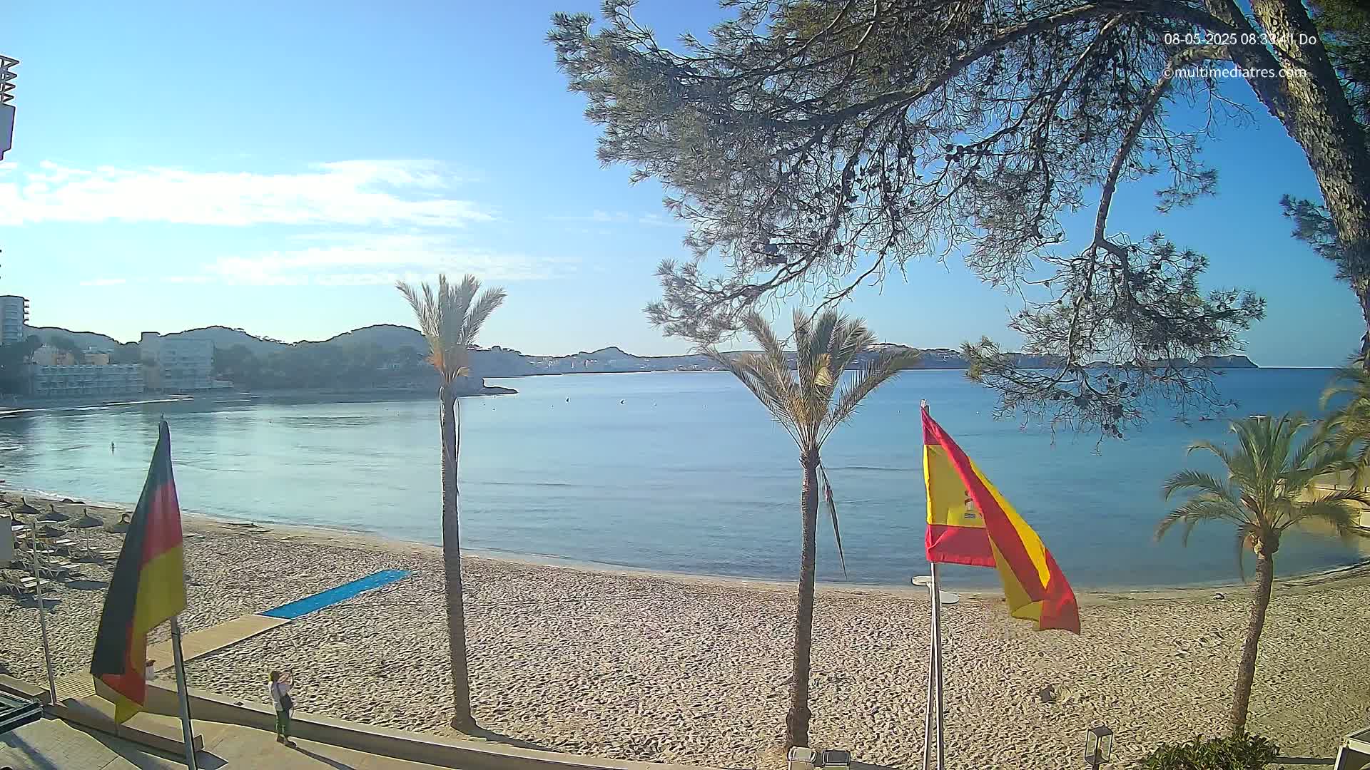 A sandy beach with calm, blue water and palm trees under a mostly sunny sky is viewed from a boardwalk with flags of Germany and Spain.
