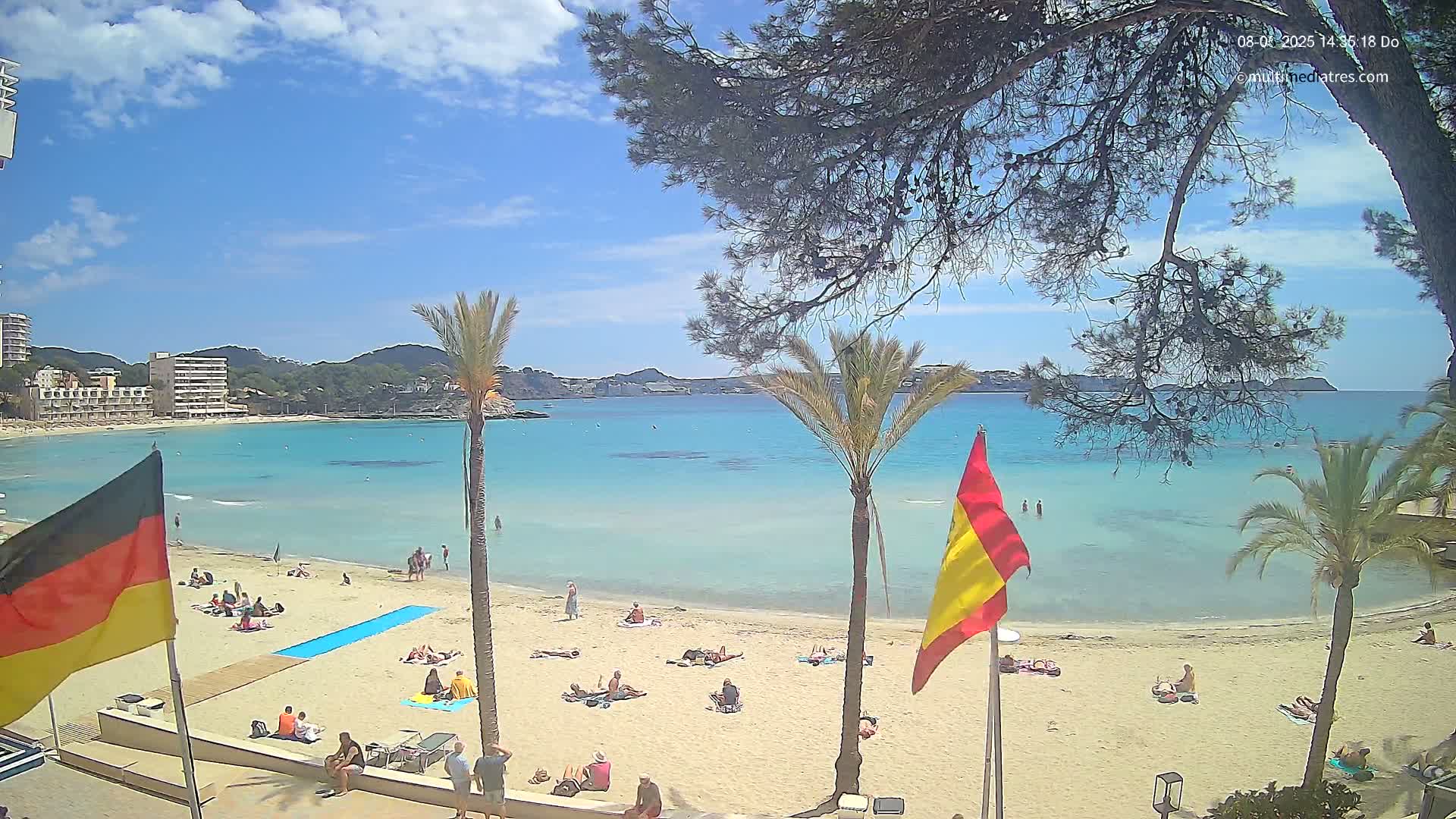 A sandy beach with scattered sunbathers is bordered by turquoise water, palm trees, and buildings under a mostly sunny sky.