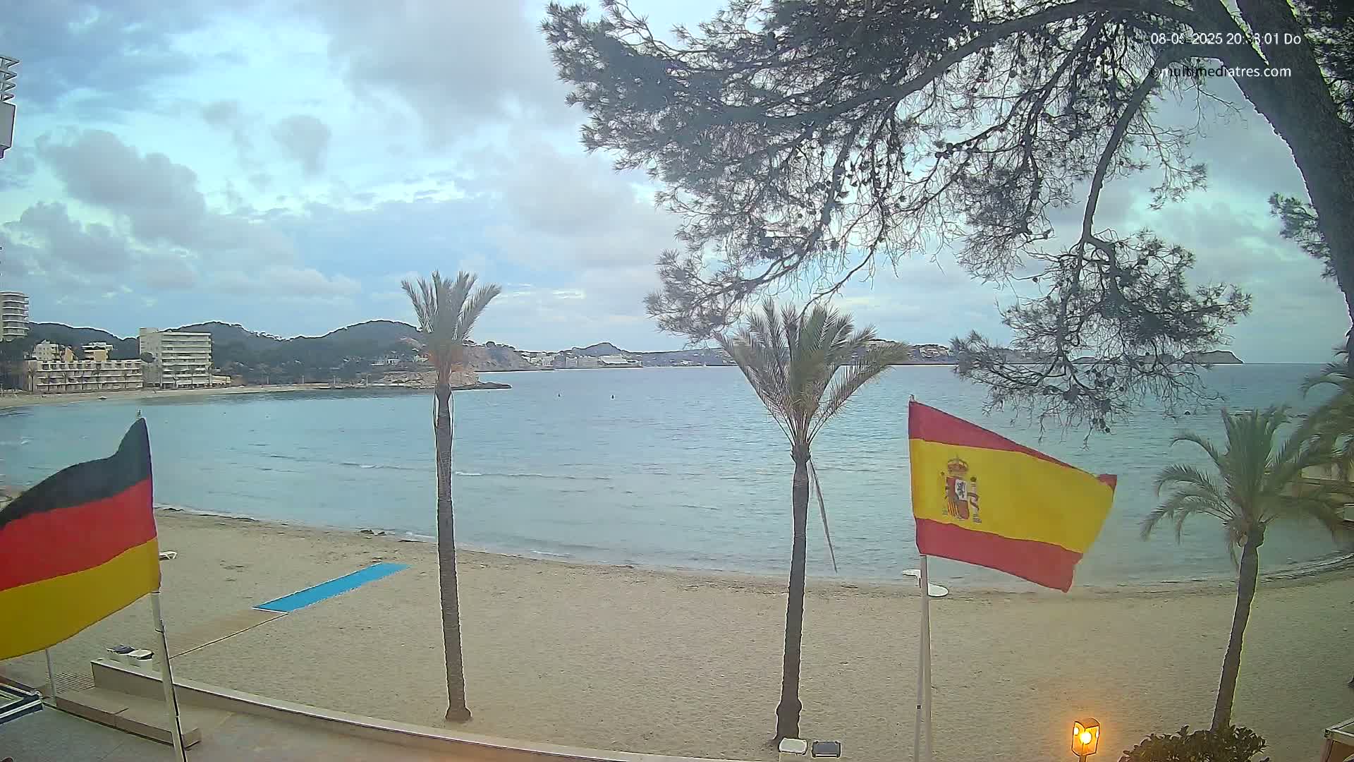 A partly cloudy day reveals a sandy beach, calm blue water, palm trees, and flags of Germany and Spain, with buildings visible in the distance.