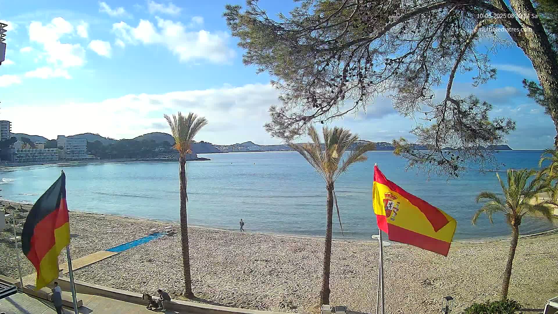 A sandy beach with calm, blue water and palm trees under a partly cloudy sky, with a German and a Spanish flag on poles.
