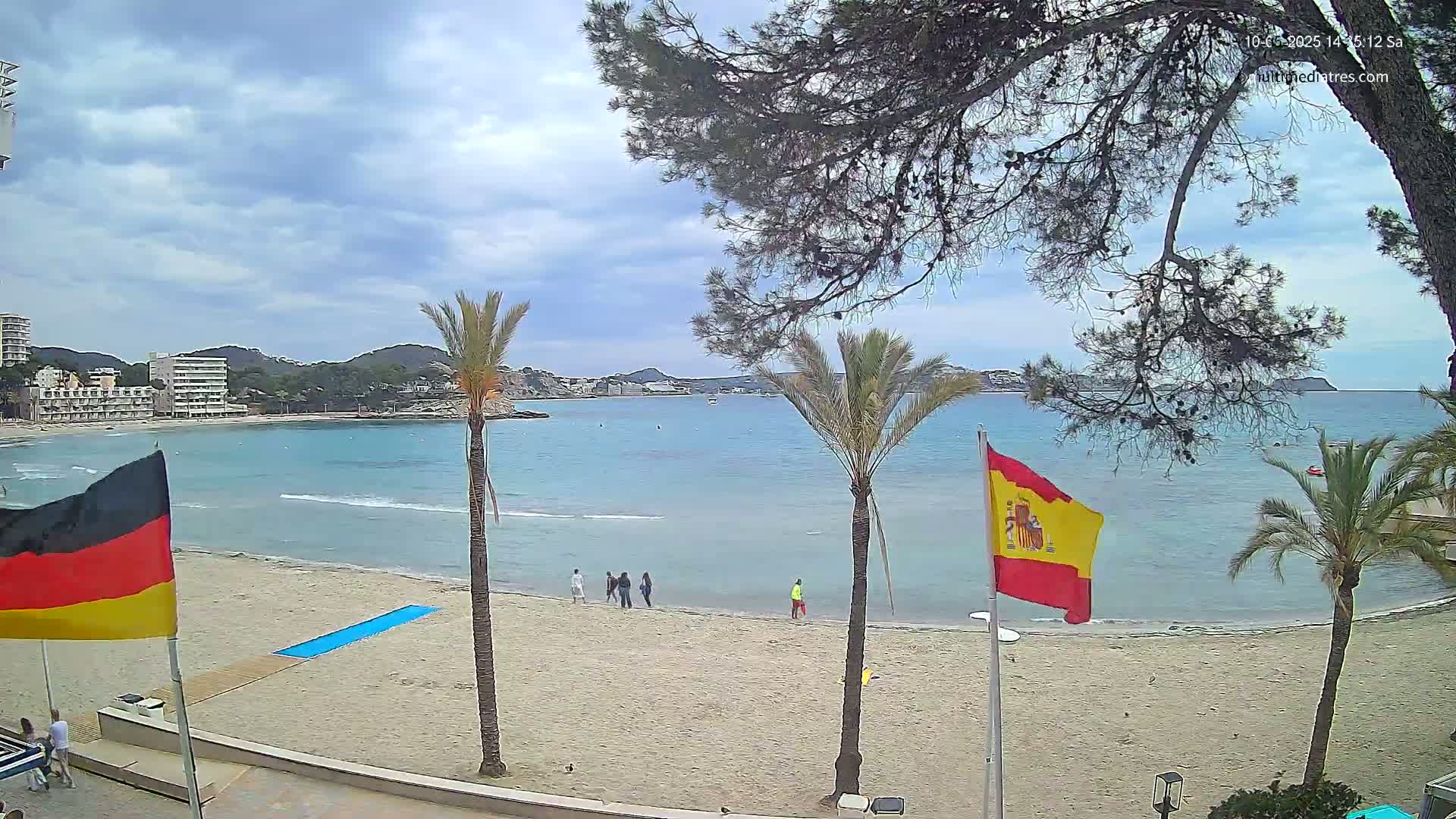 A sandy beach with calm, light-blue water, palm trees, and a few people are visible under a partly cloudy sky.