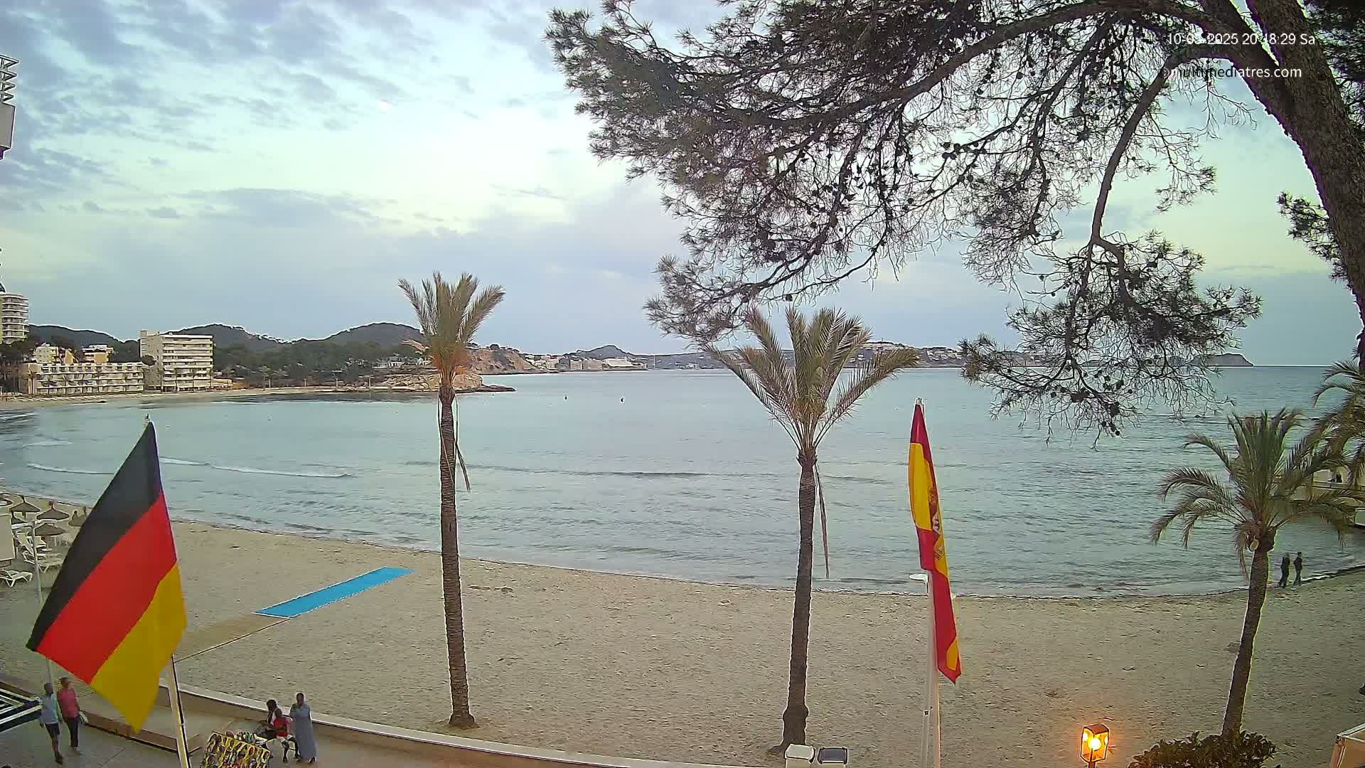 A sandy beach with calm, blue water, palm trees, and flags overlooks a bay with buildings and hills in the distance under a mostly cloudy sky.