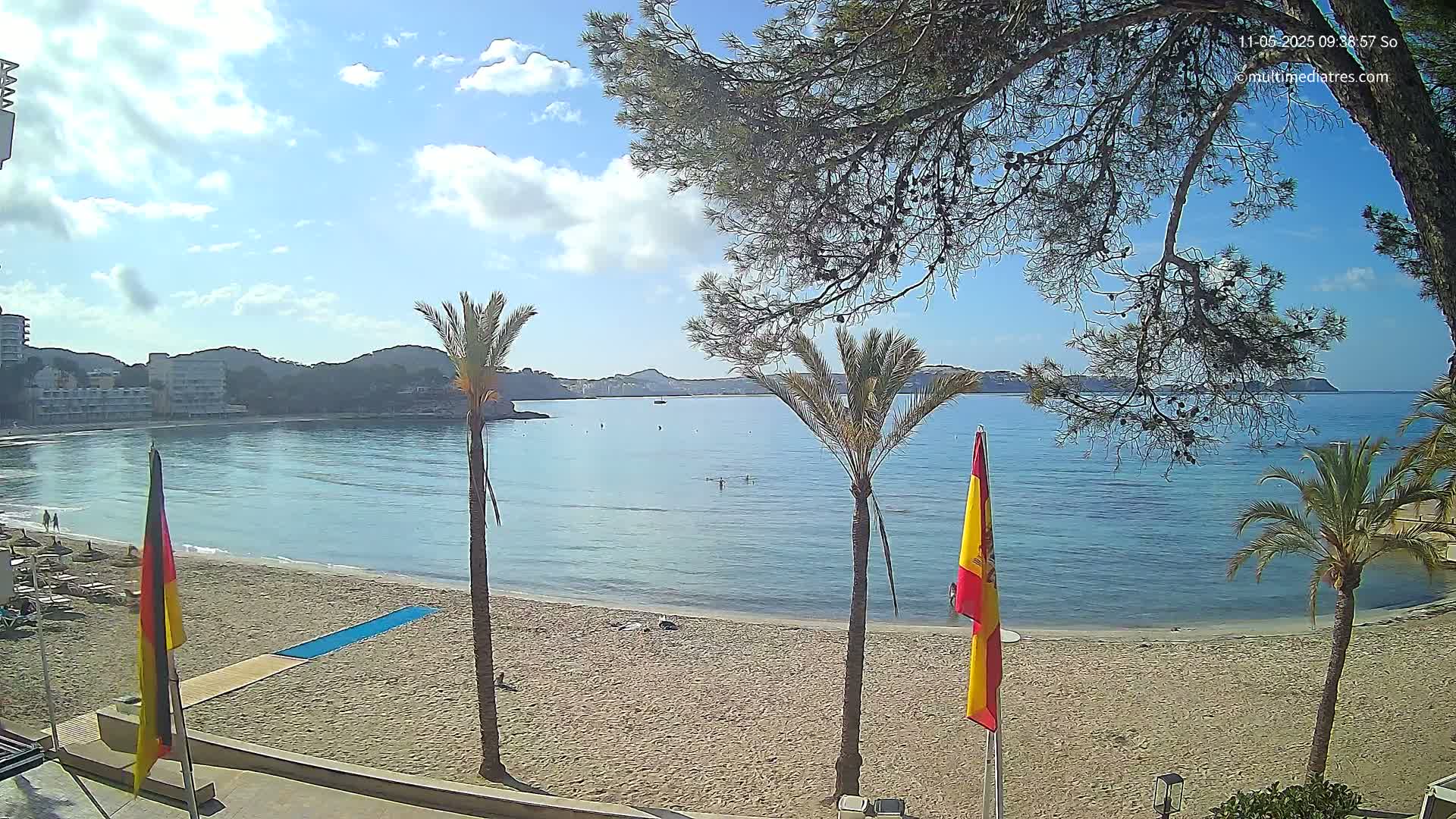 A sandy beach with palm trees and calm, clear blue water stretches out to a bay bordered by hills under a mostly sunny sky.
