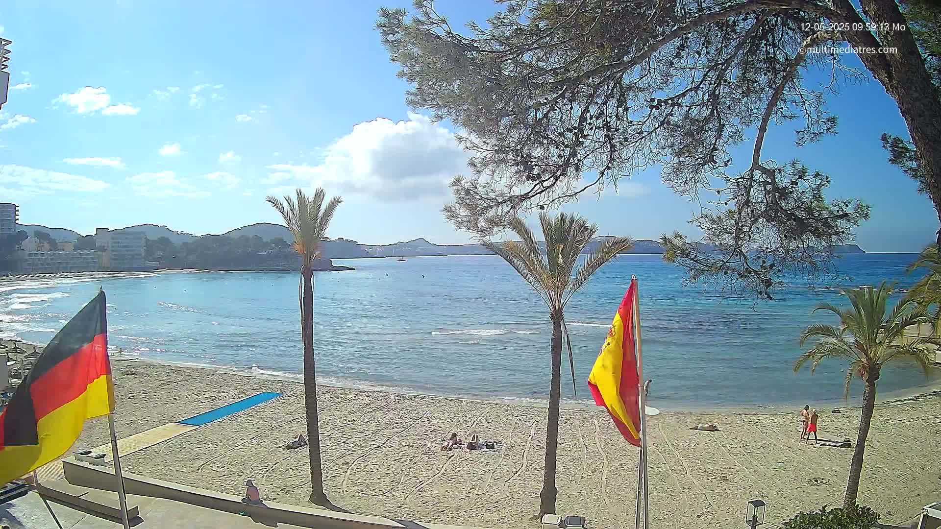 A sandy beach with calm, blue water and a few people sunbathing, under a mostly sunny sky, is framed by palm trees and buildings in the background.