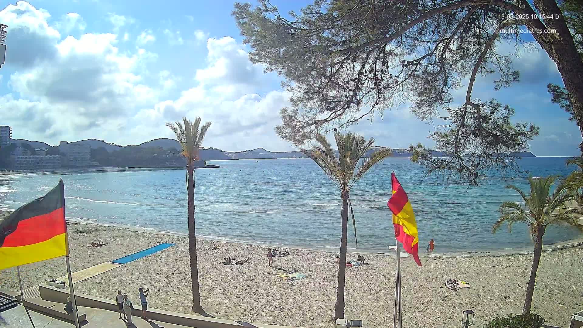 A sandy beach with calm, light-blue water, palm trees, and a few people relaxing under a partly cloudy sky.