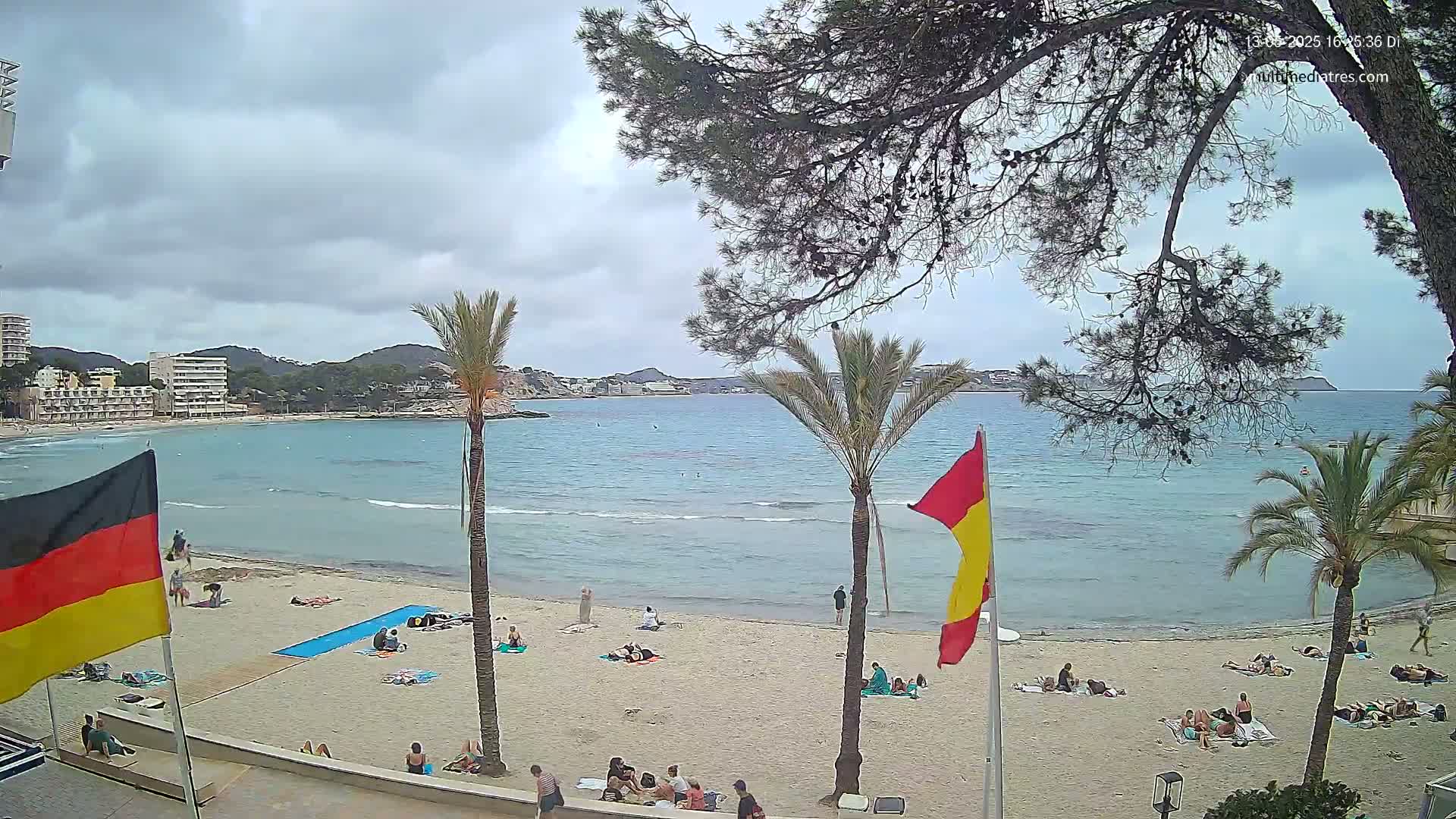 A sandy beach with scattered sunbathers is visible beneath a cloudy sky, overlooking a calm, turquoise bay framed by palm trees and buildings.