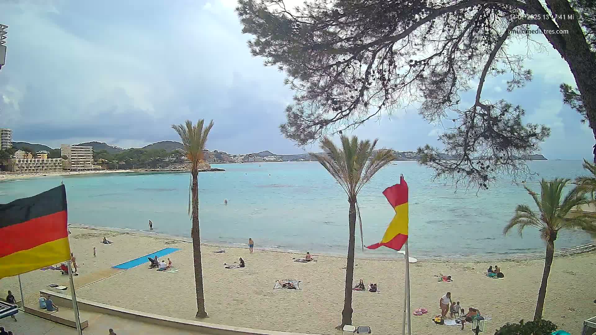 A partly cloudy day at a sandy beach with calm, light-blue water, palm trees, and a few people scattered on the sand and in the water, with flags planted in the sand.
