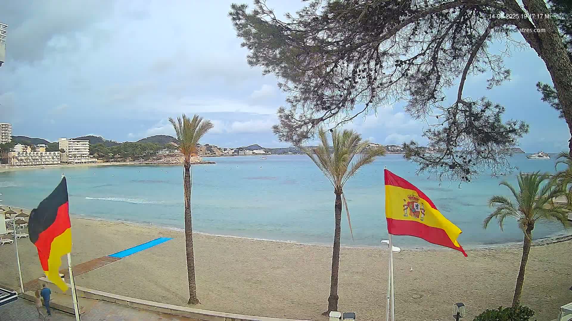 A sandy beach with calm, light blue water is flanked by palm trees and buildings under an overcast sky, with German and Spanish flags displayed nearby.