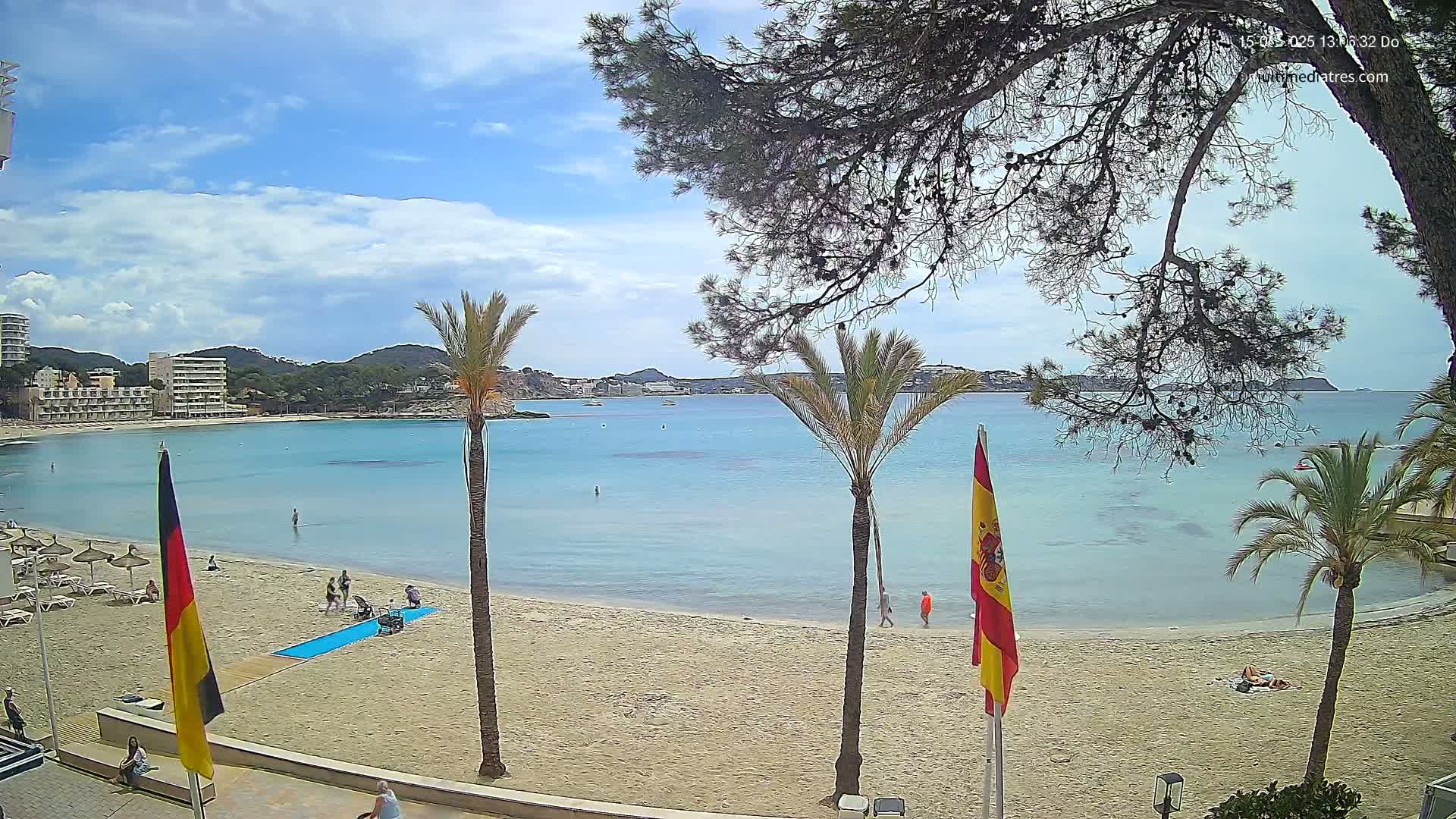 A sandy beach with calm, light-blue water, palm trees, and a few people is visible under a partly cloudy sky.