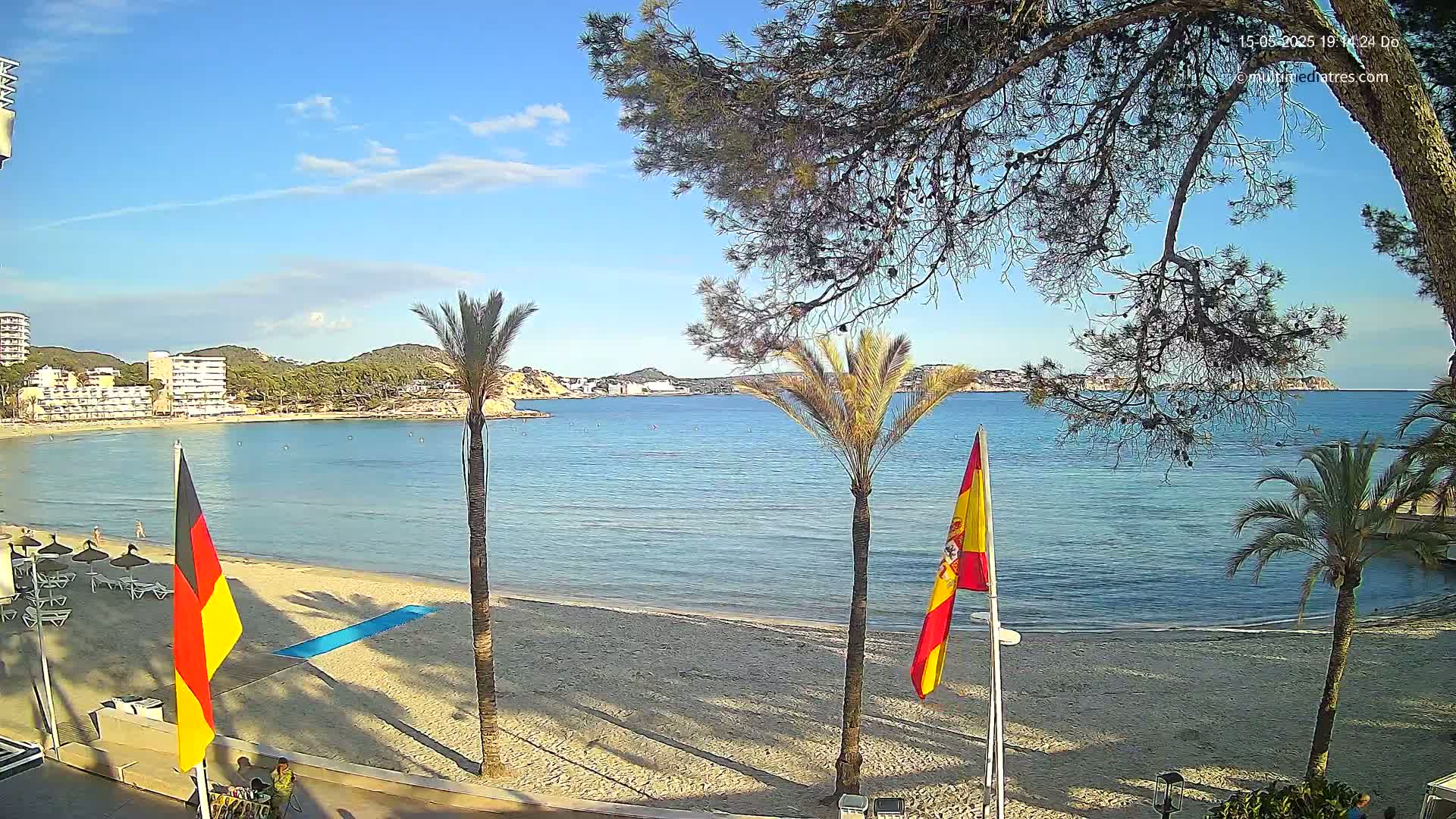 A sandy beach with calm, clear blue water, palm trees, and flags is visible under a mostly sunny sky.