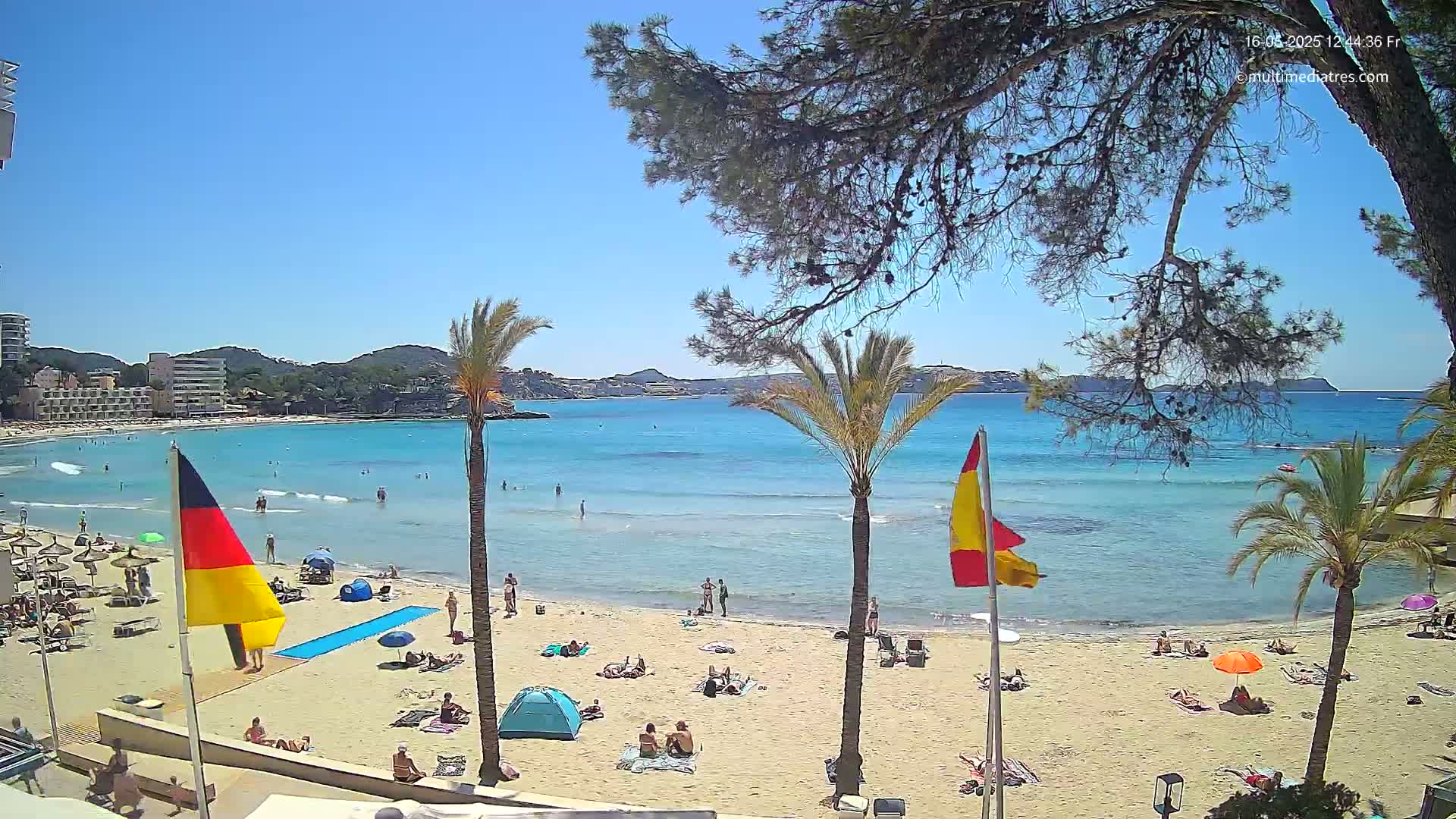 A sunny beach scene shows a sandy shore with people relaxing, umbrellas, and flags, all overlooking calm, turquoise water under a clear blue sky.