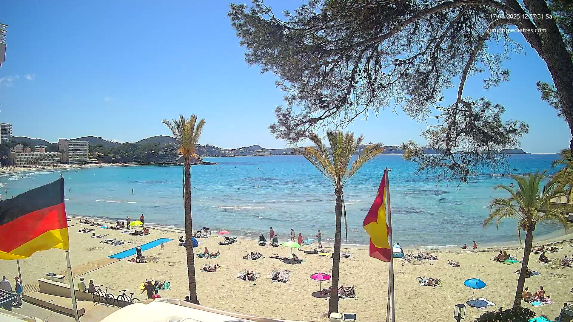 A sandy beach with scattered sunbathers is shown, under a clear blue sky, next to a calm, turquoise sea with distant hills and a few palm trees.