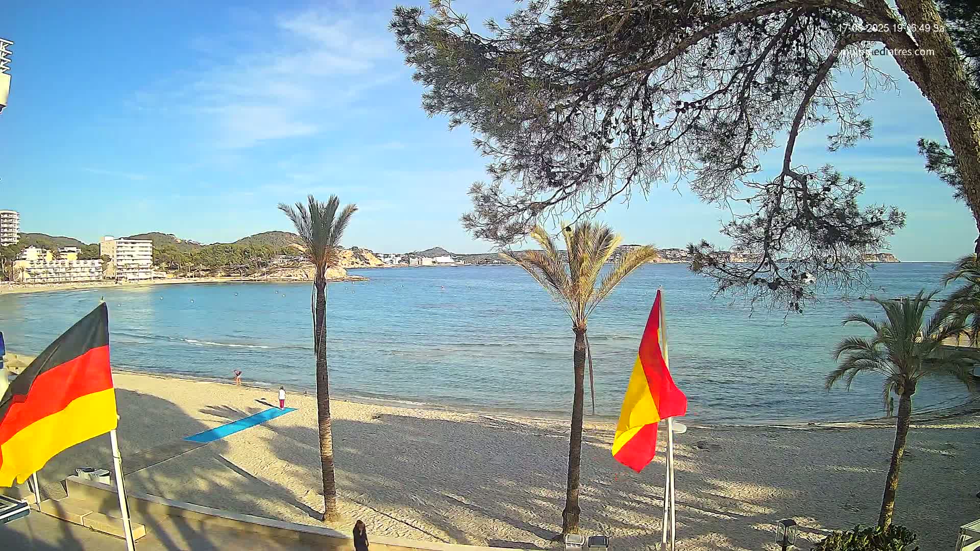 A sunny beach scene shows a calm sea with palm trees and flags on a sandy shore, overlooking buildings and hills in the distance.