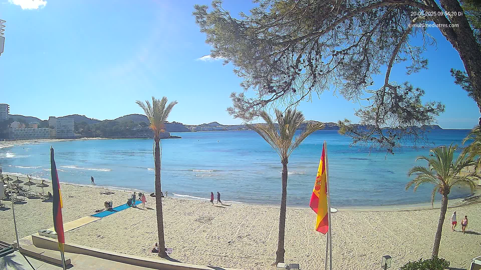 A sandy beach with palm trees and a few people is seen, under a sunny sky, overlooking a calm, light-blue bay and distant hills.