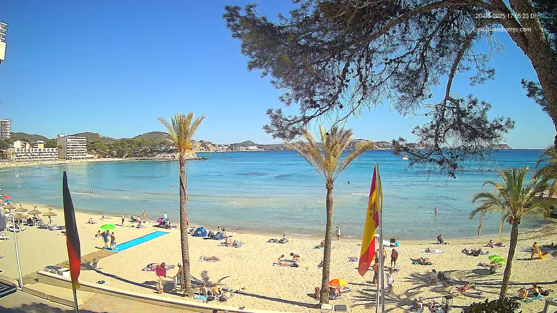 A sunny beach scene shows a calm, turquoise bay with a sandy shore dotted with sunbathers, palm trees, and flags.