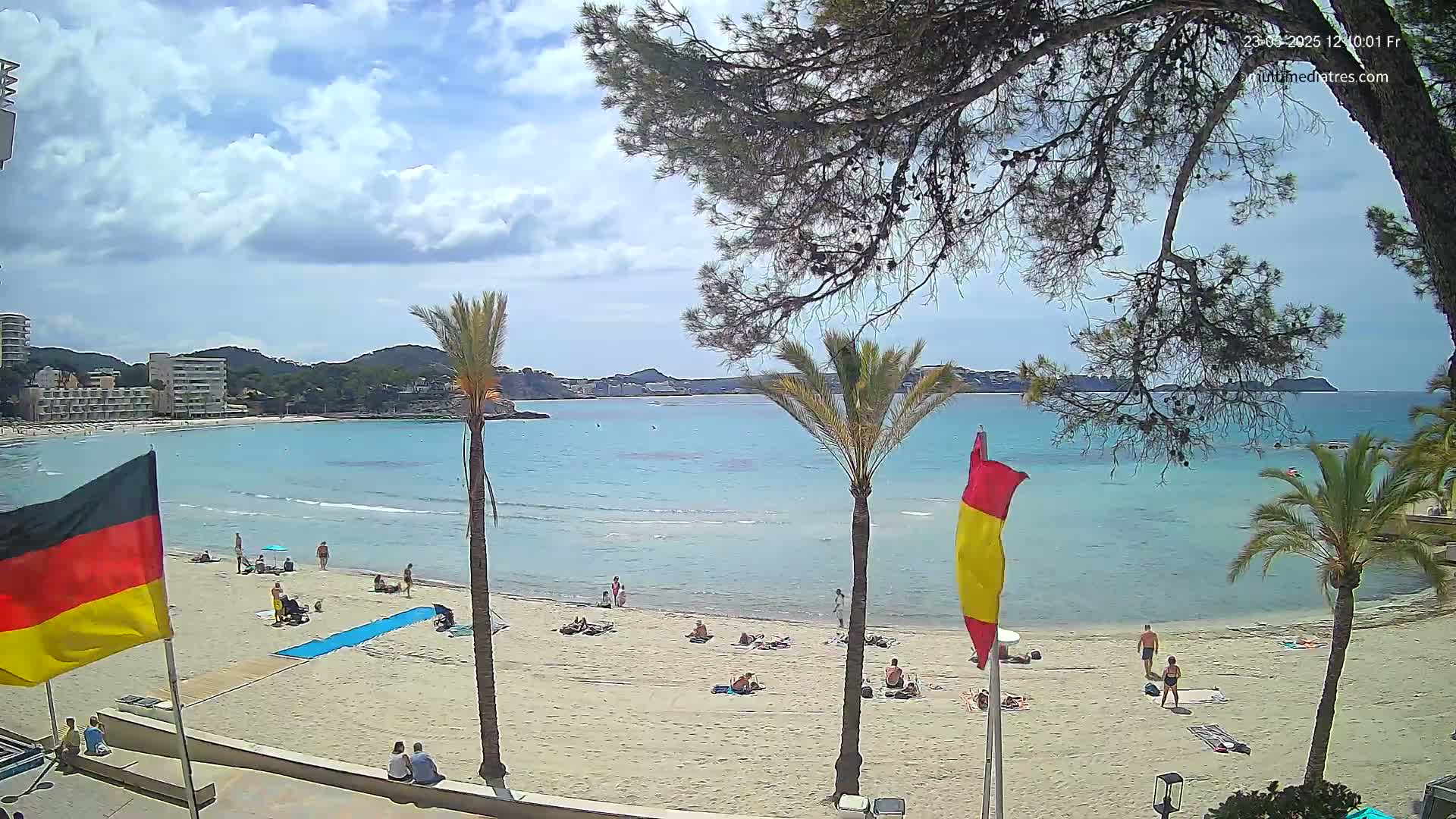 A sandy beach with people sunbathing and swimming in clear, turquoise water under a partly cloudy sky, palm trees and other vegetation lining the beach.