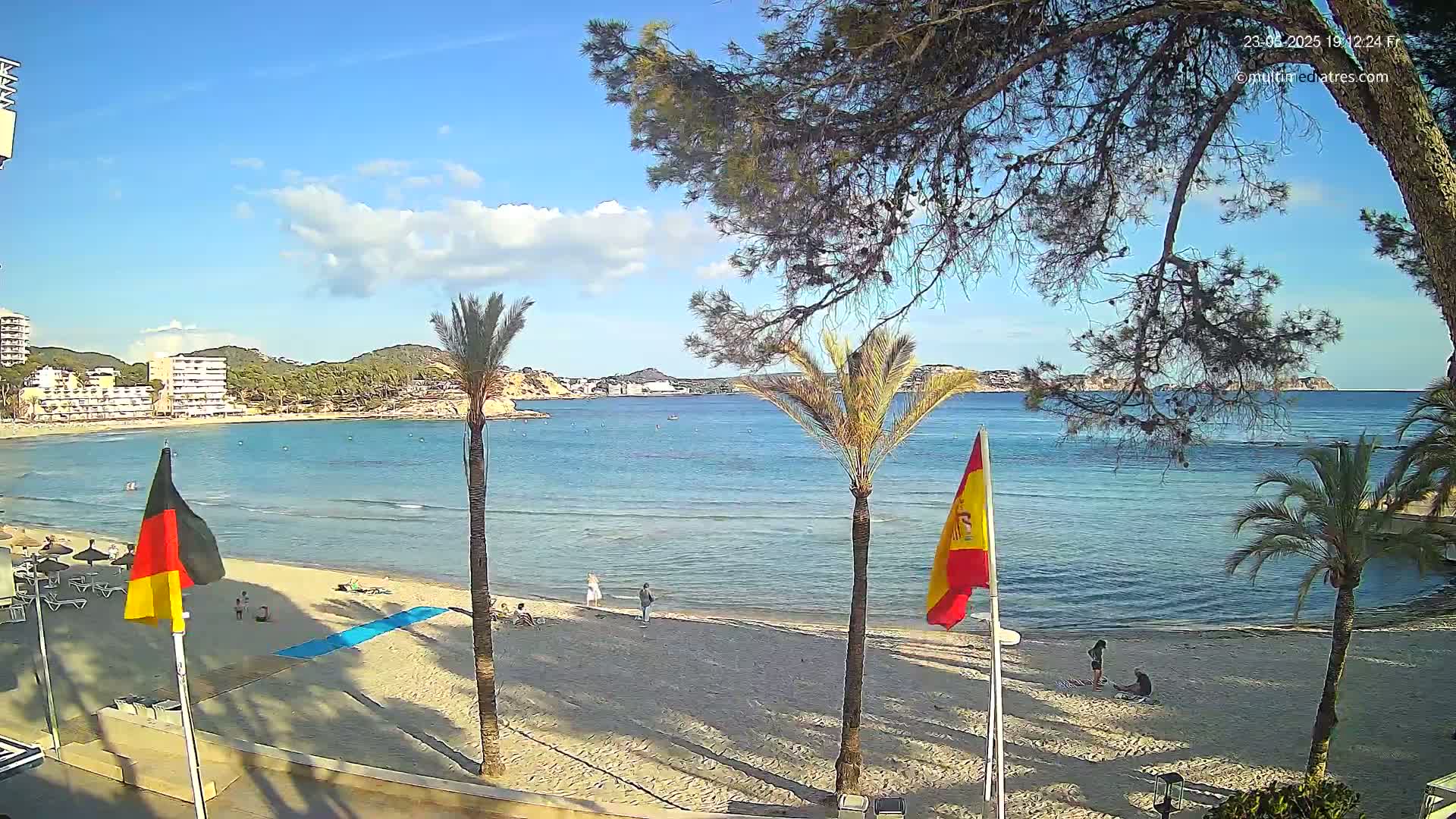 A sandy beach with palm trees and flags is visible, overlooking a calm, blue bay under a partly cloudy sky.