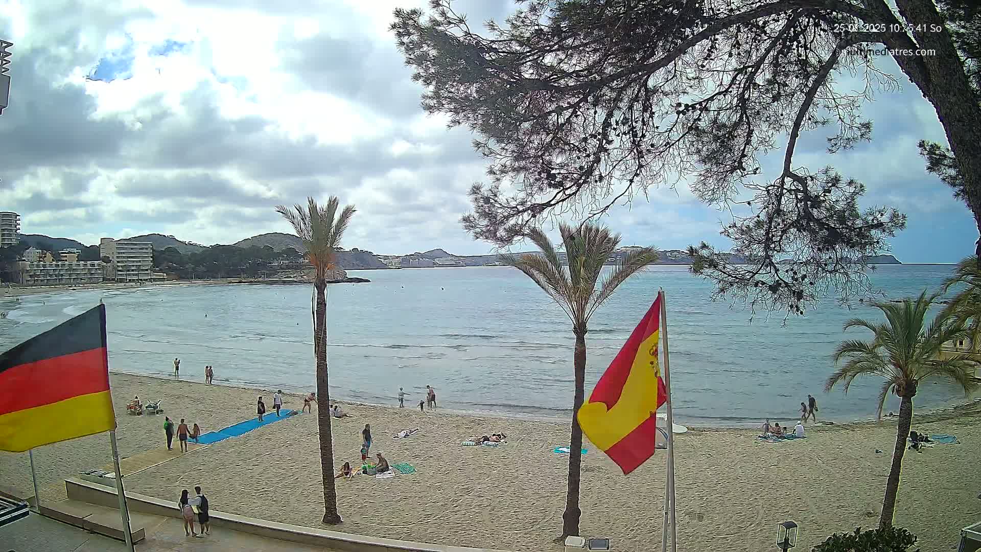 A partly cloudy day at a sandy beach with calm water, palm trees, buildings in the distance, and a few people scattered on the sand.