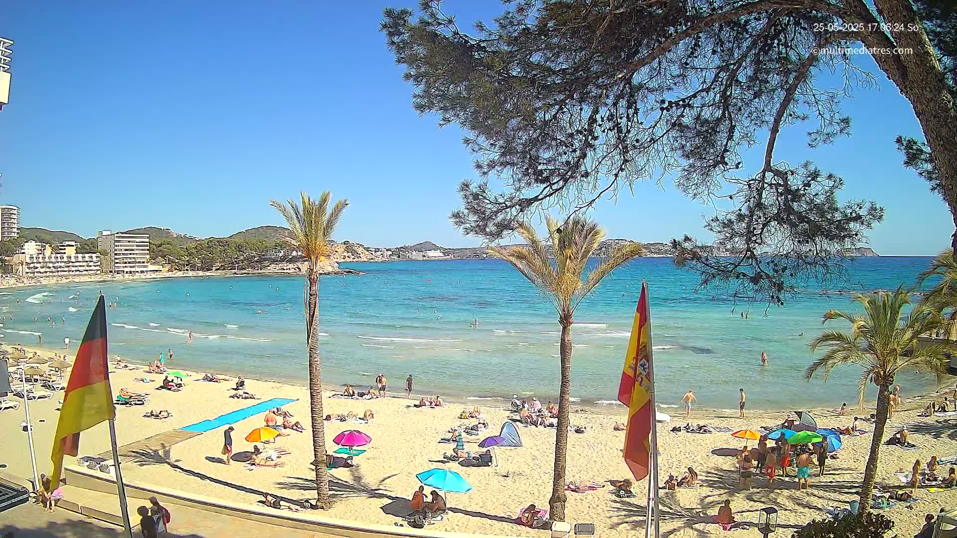 A sunny beach scene shows a sandy shore with many people relaxing under umbrellas, palm trees, and clear, turquoise water extending to a distant coastline.