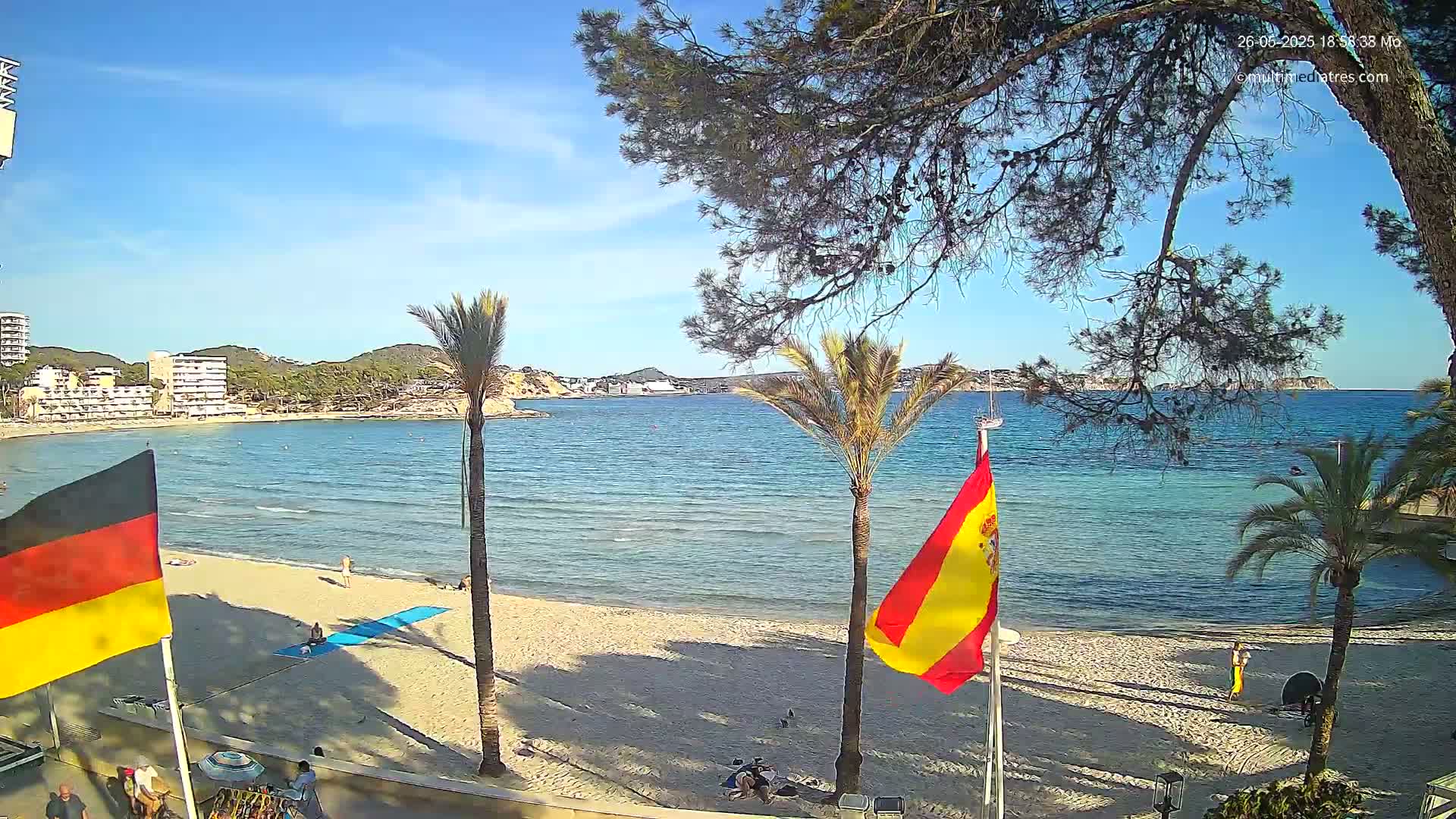 A sandy beach with calm blue water, palm trees, and flags of Germany and Spain is visible under a sunny sky.