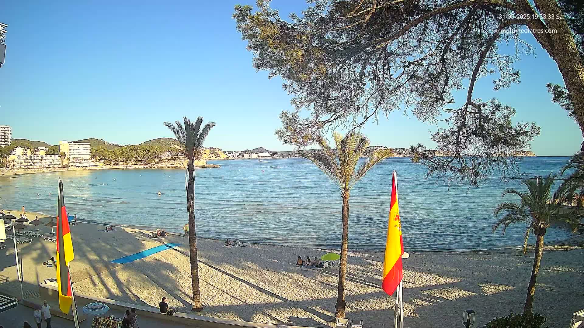 A sandy beach with calm, clear water, palm trees, and a few people are visible under a clear blue sky.