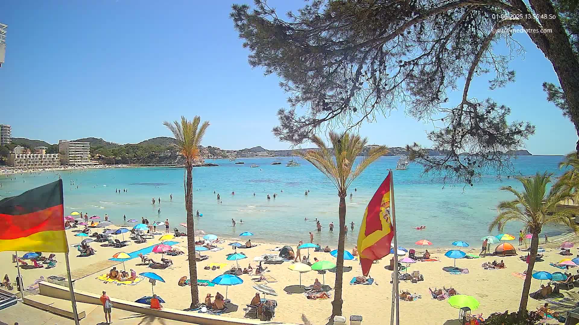 A sunny beach scene shows a sandy shore crowded with people under colorful umbrellas, calm turquoise water with swimmers, and boats in the distance, all under a bright blue sky.