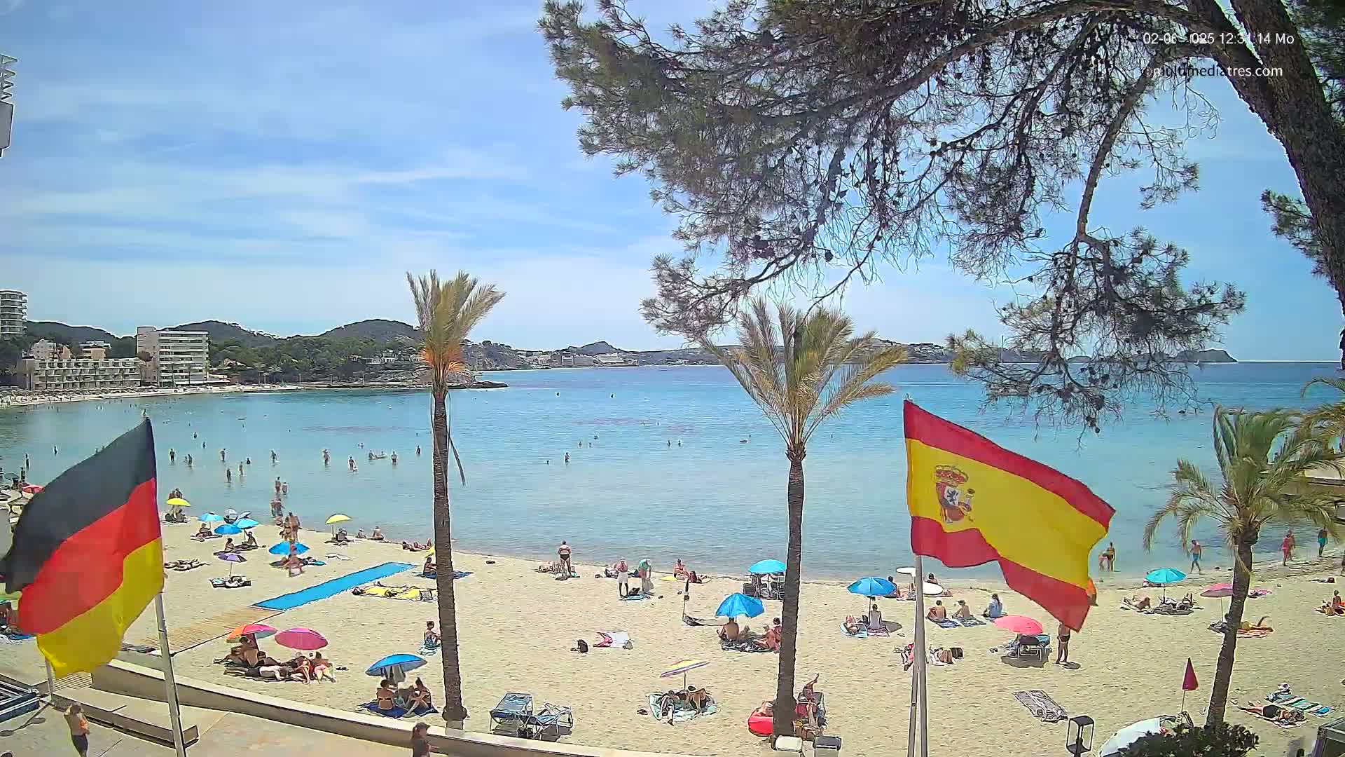 A sunny beach scene shows a sandy shoreline with many people relaxing under umbrellas, swimming in calm, clear water, and a few flags planted in the sand.