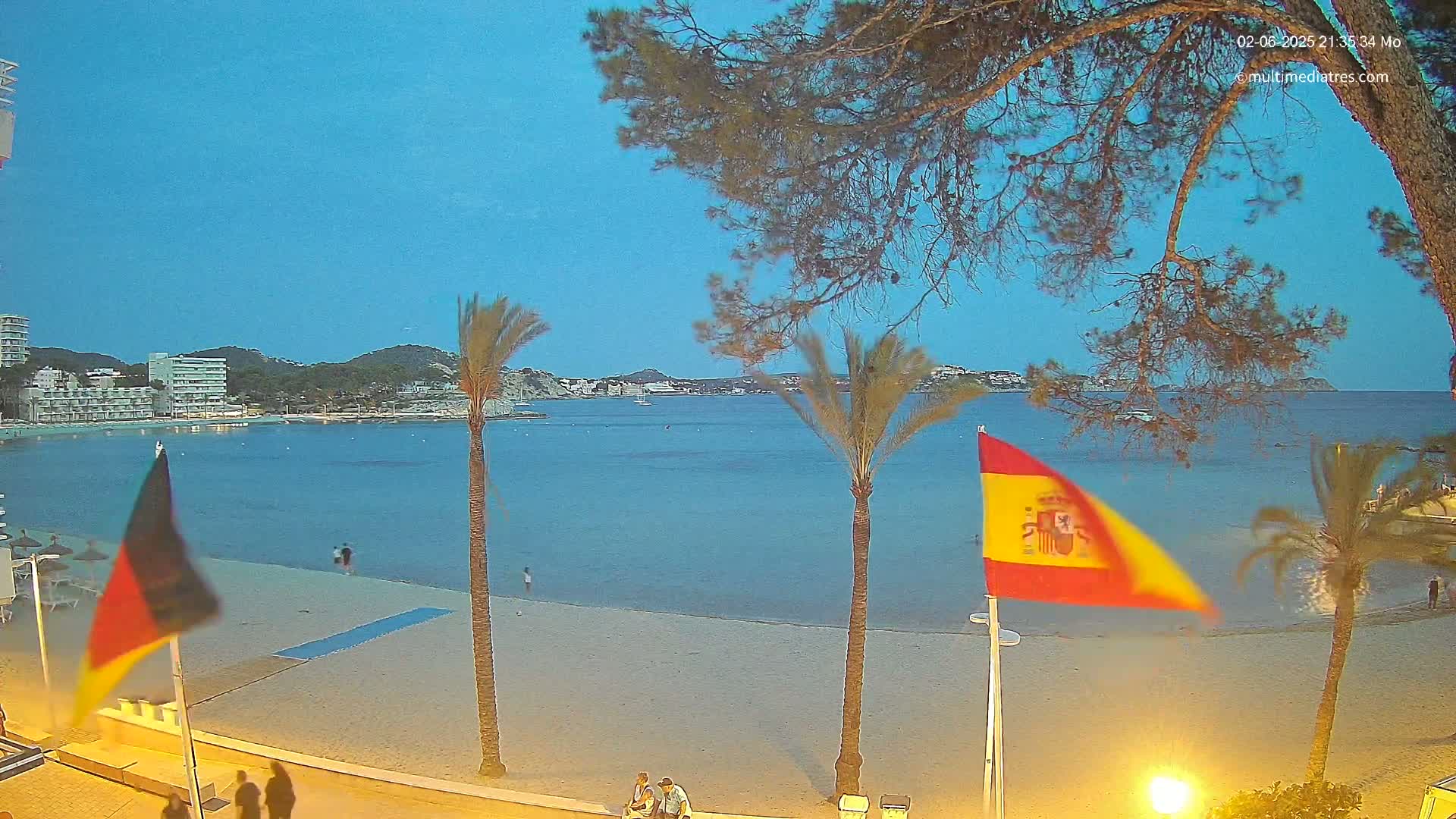 A sandy beach with calm blue water, palm trees, and flags is illuminated at dusk under a clear sky.