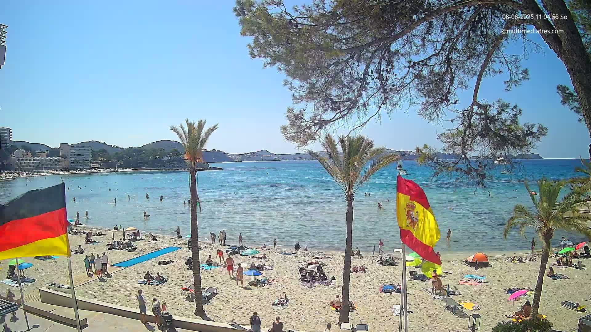 A sunny beach scene shows a sandy shore with bathers and beach umbrellas, calm turquoise water, palm trees, and buildings in the background.
