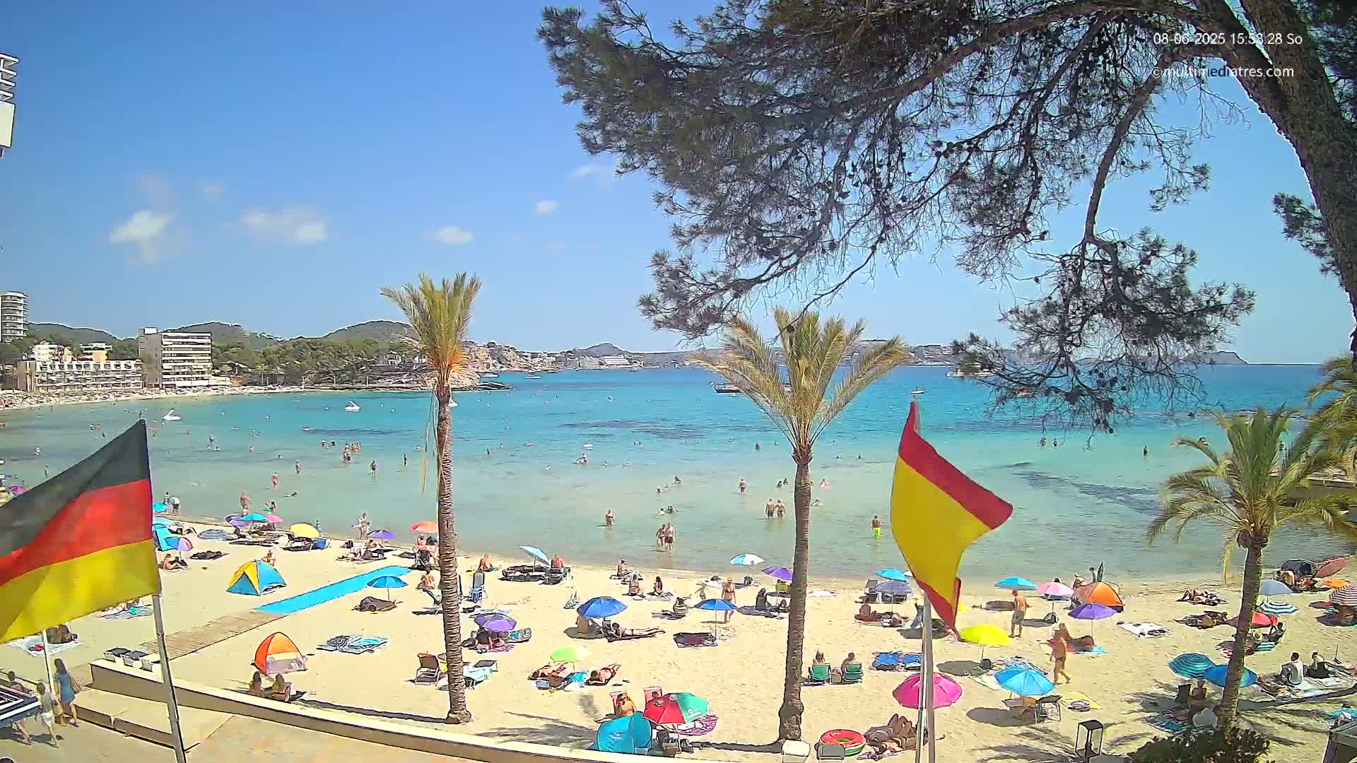 A sunny beach scene shows a sandy shore with numerous beach umbrellas and bathers in shallow, clear turquoise water, backed by buildings and hills under a mostly clear sky.