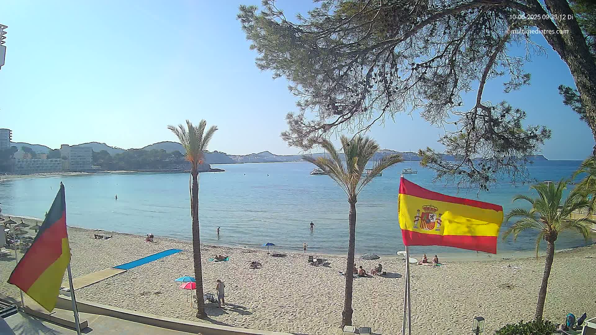 A sunny beach scene shows calm, clear blue water, sandy shores with people relaxing, palm trees, and flags of Germany and Spain waving in the breeze.