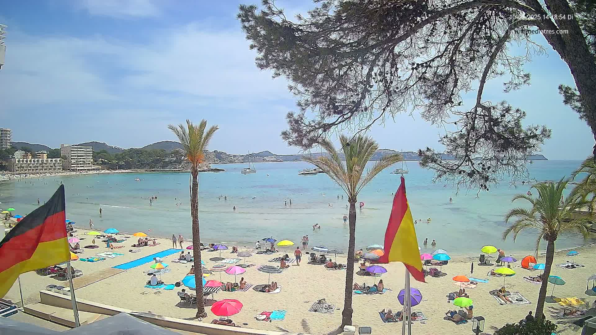 A sunny beach scene with calm, clear water, numerous colorful umbrellas, and people relaxing on the sand, backed by palm trees and buildings.