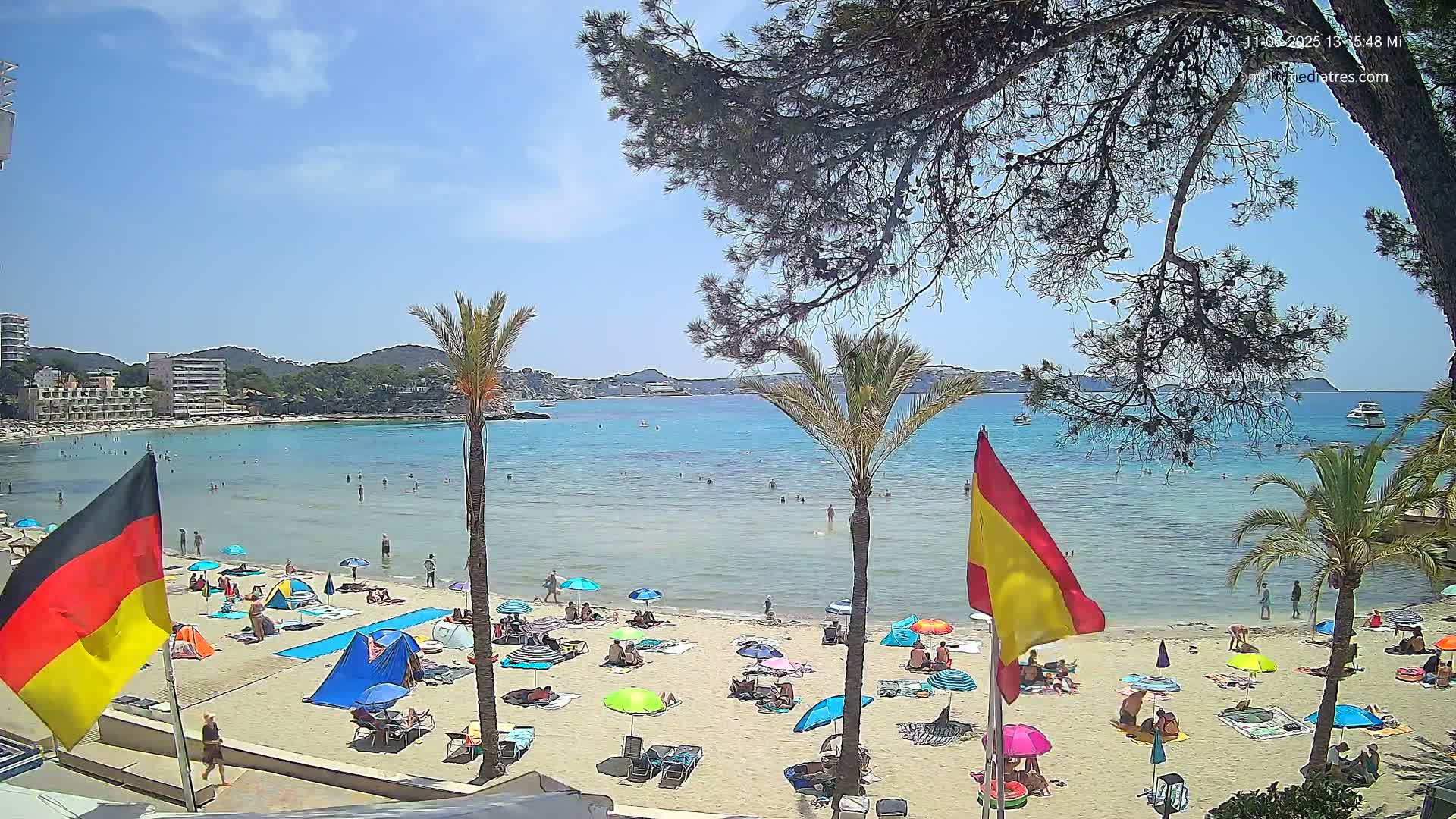 A sunny beach scene shows a calm, turquoise bay with people swimming and sunbathing under umbrellas, palm trees, and flags on the sand.