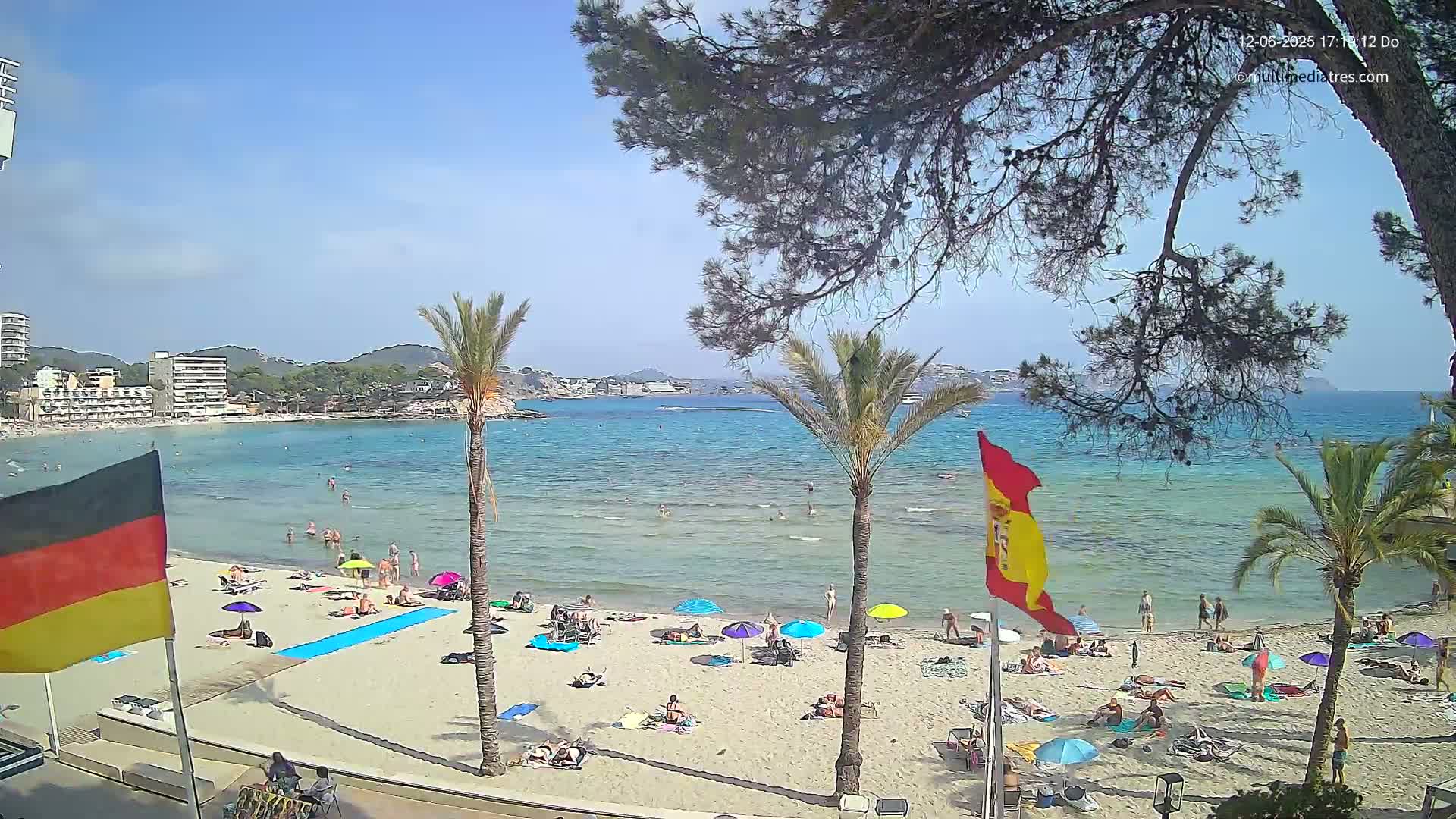 A sunny beach scene shows a sandy shore with numerous beachgoers under umbrellas, palm trees, and flags, overlooking calm, clear turquoise water and distant hills.