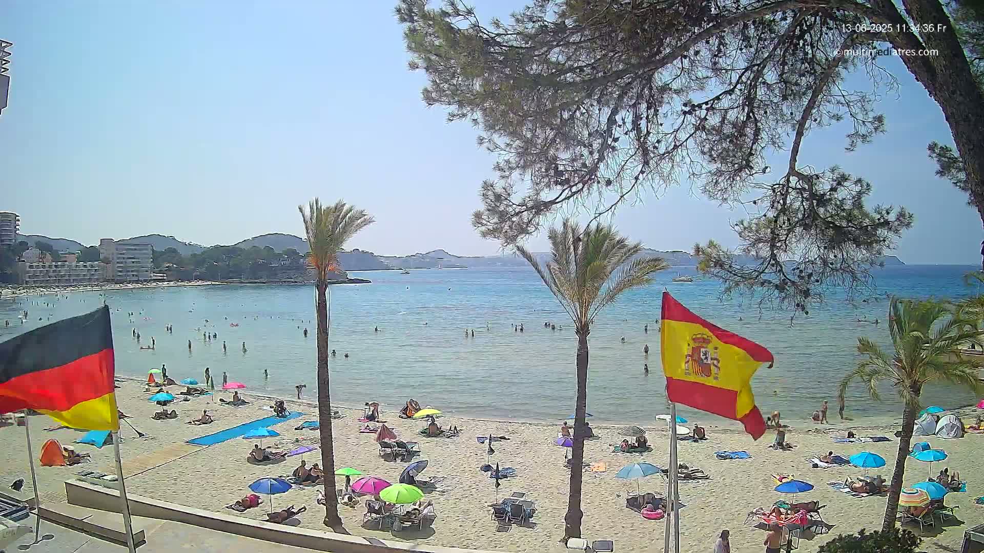 A sunny beach scene shows a calm, clear body of water with people swimming and sunbathing, palm trees lining the shore, and flags of Germany and Spain displayed.