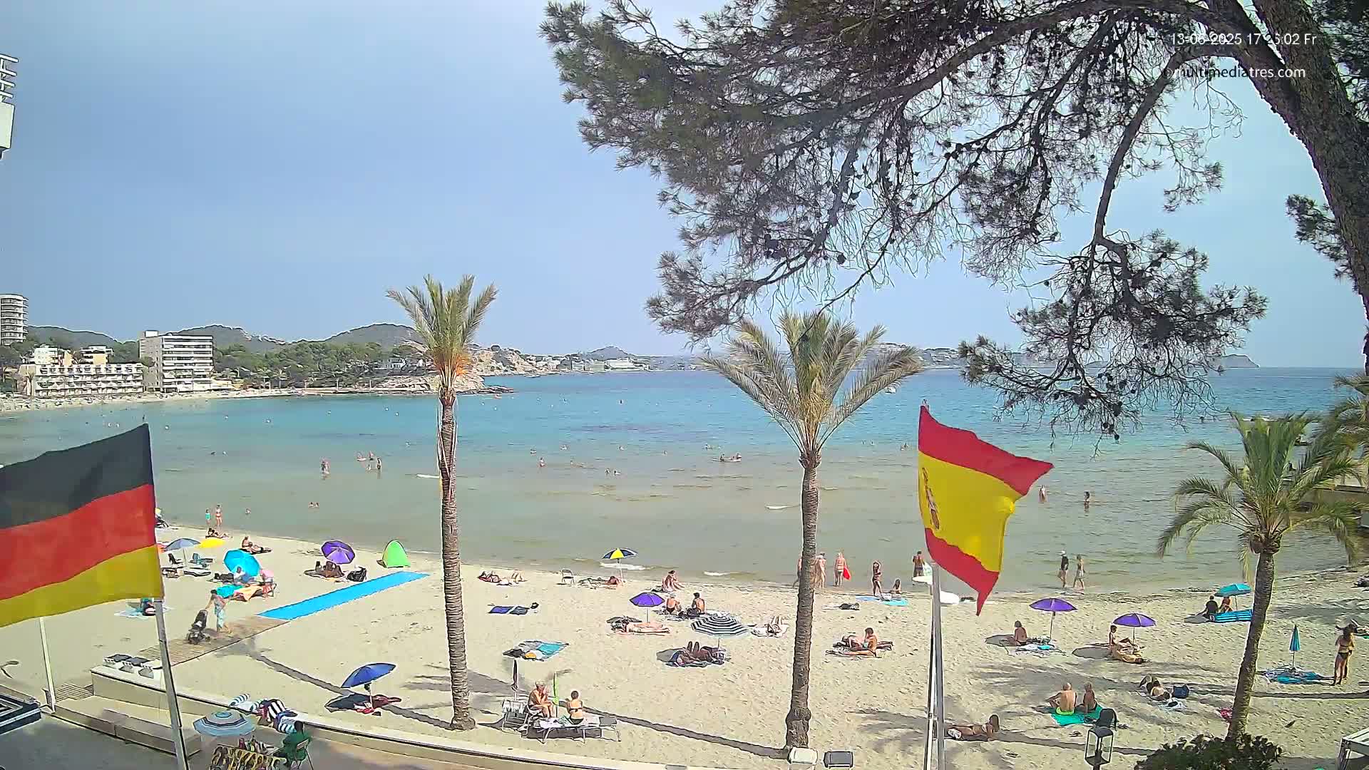 A sandy beach with scattered sunbathers and palm trees, overlooks a calm, turquoise sea under a clear, sunny sky.