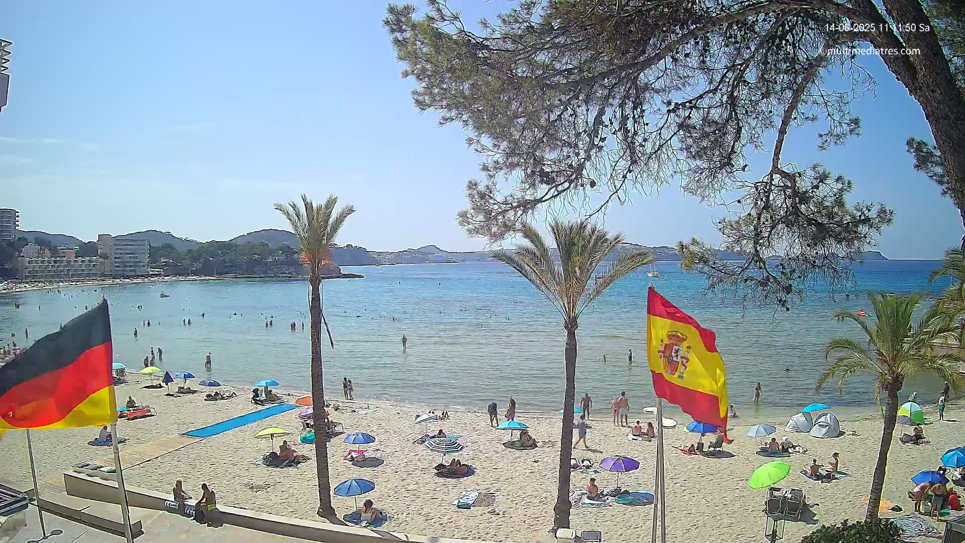 A sunny beach scene shows a calm, turquoise bay with people swimming and sunbathing under colorful umbrellas, palm trees, and flags of Germany and Spain.