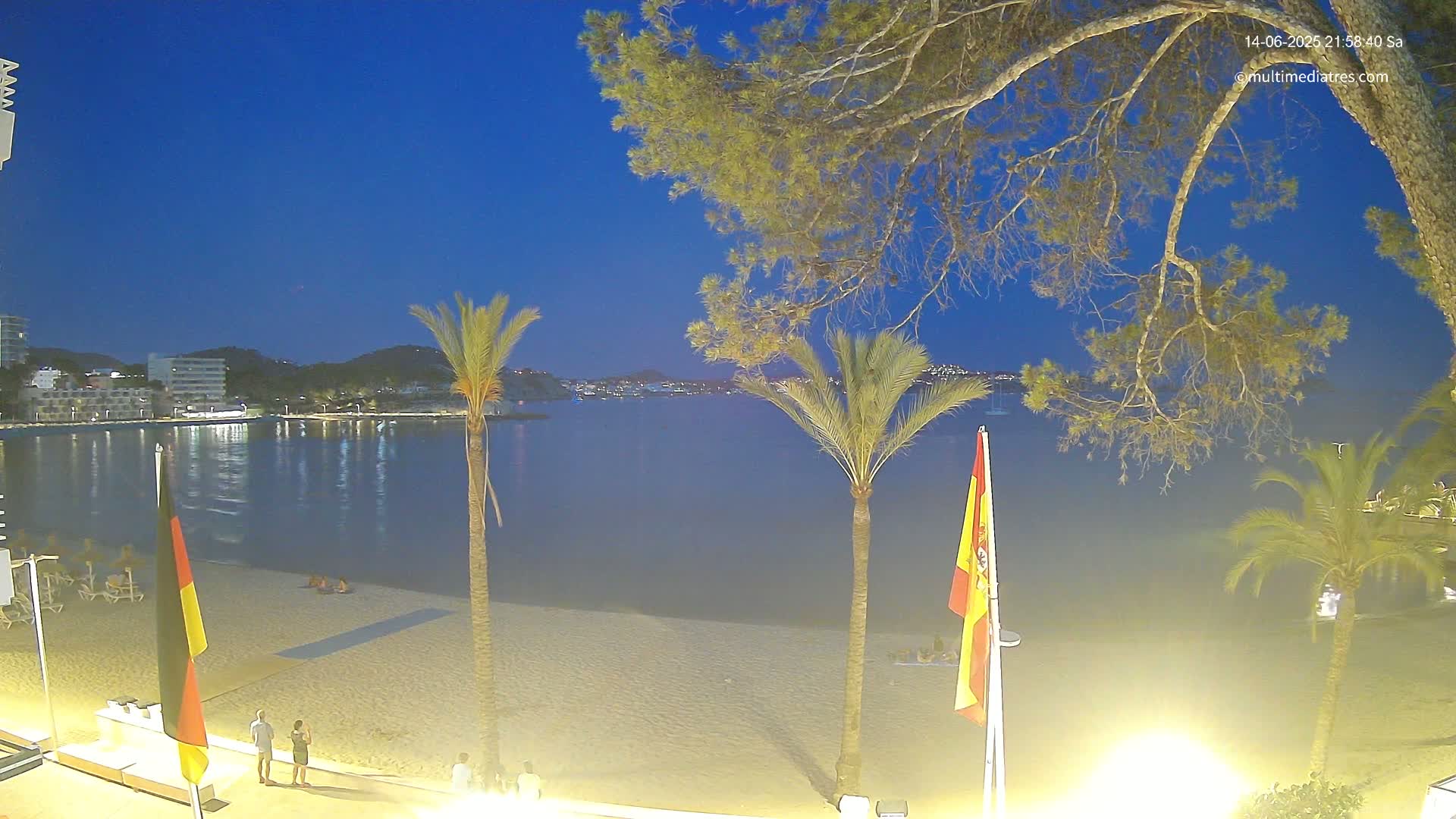 A sunny beach scene shows a sandy shore with people relaxing under colorful umbrellas, palm trees, and clear turquoise water extending to a distant coastline.