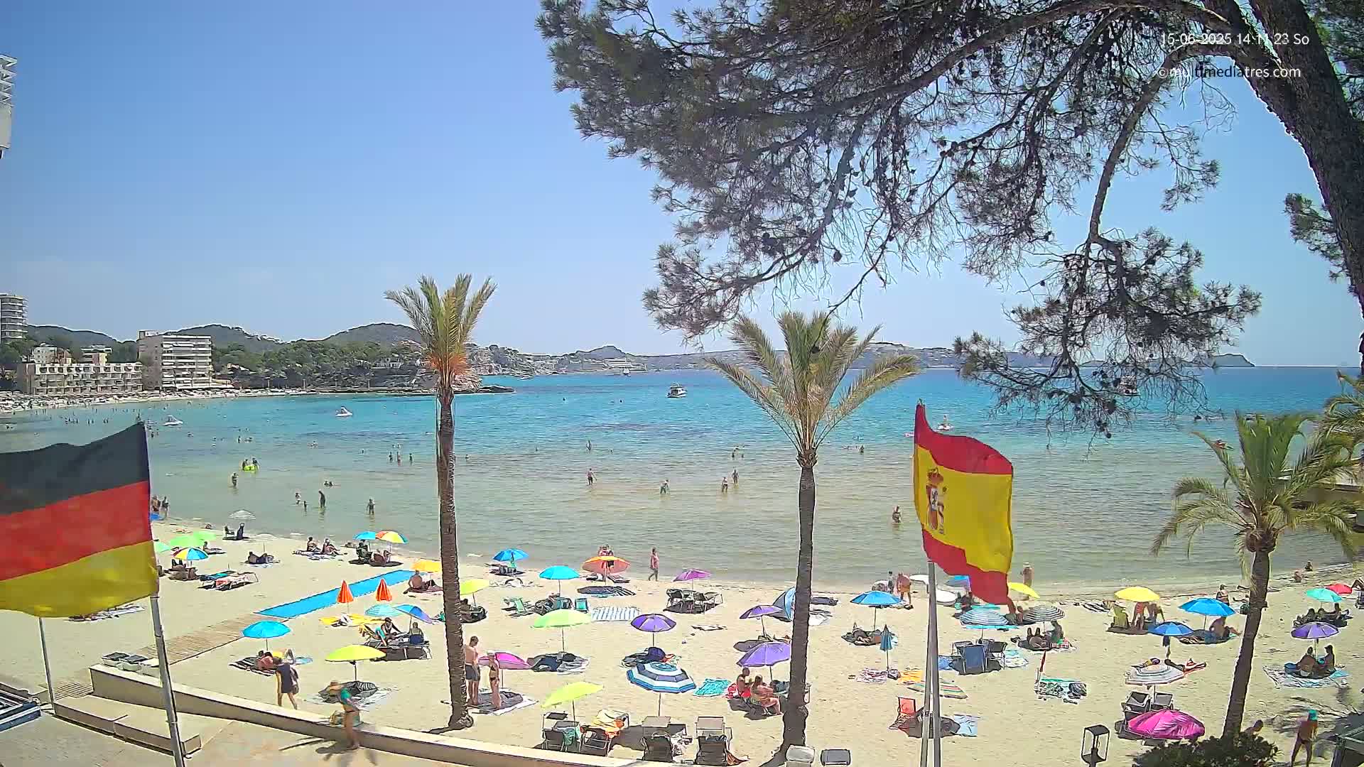 A sunny beach scene shows a sandy shore with numerous colorful umbrellas and sunbathers, palm trees, and calm turquoise water with boats in the distance.
