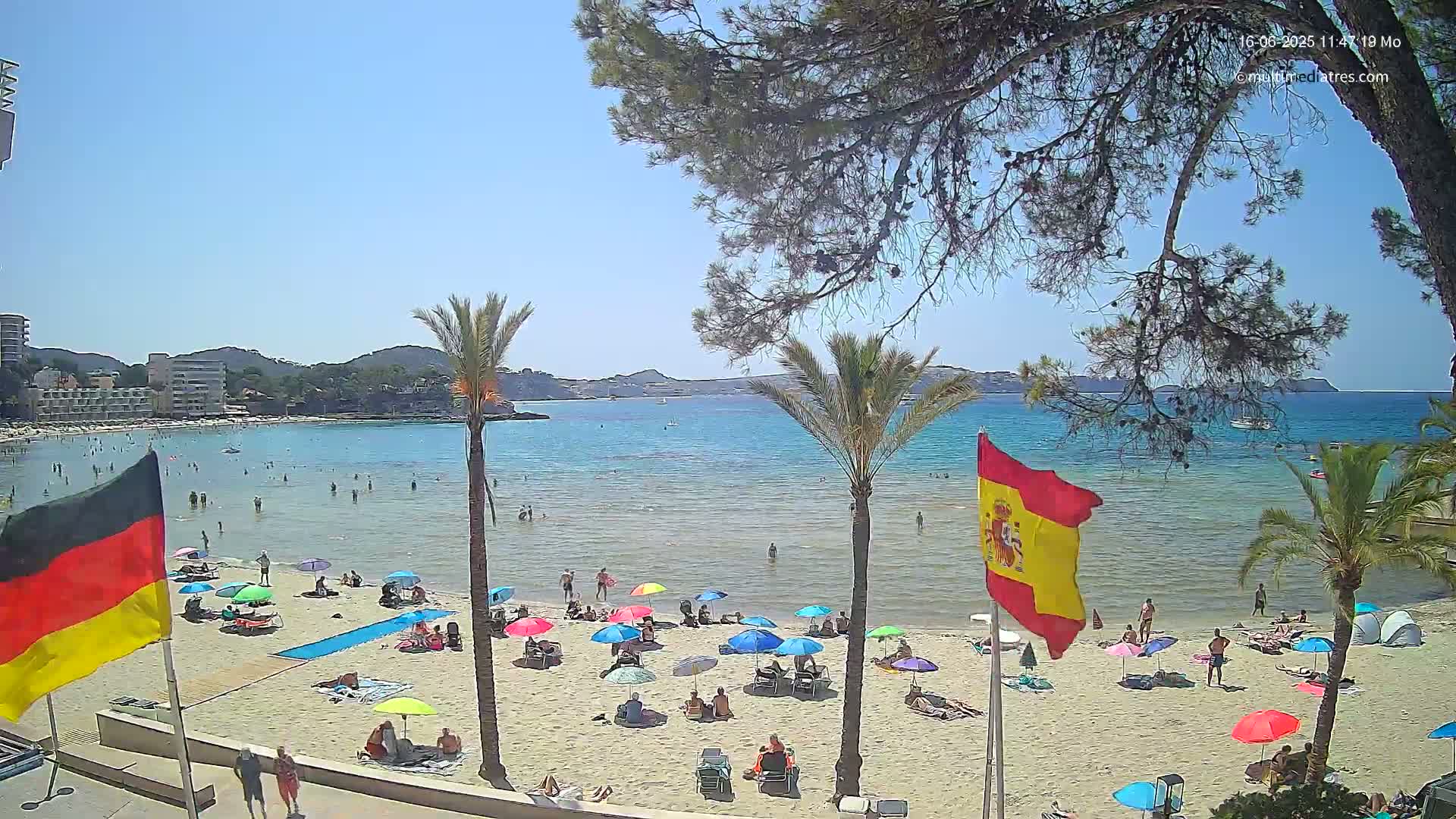 A sunny beach scene shows many people relaxing under colorful umbrellas on a sandy beach next to calm, clear blue water, with palm trees and flags in the foreground.