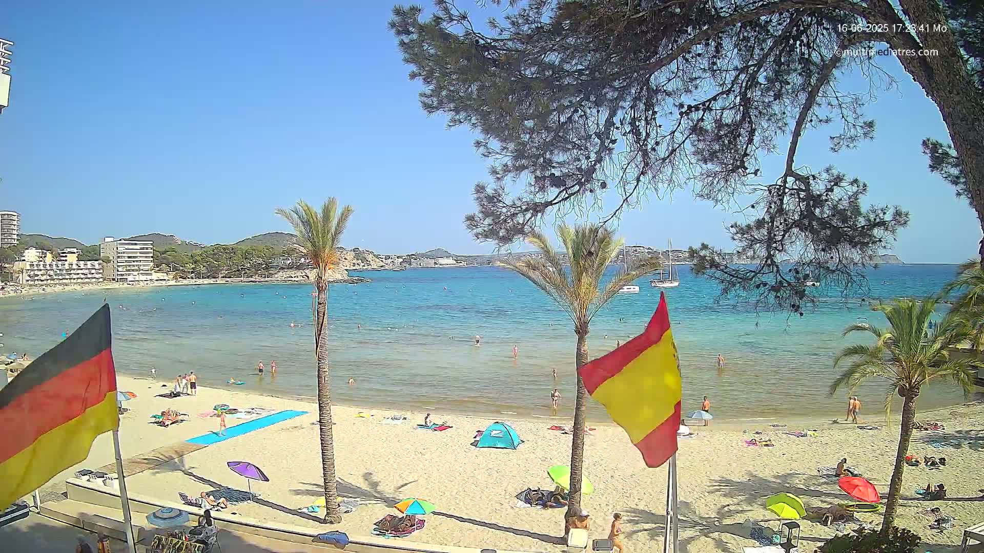 A sunny beach scene shows a sandy shore with bathers, palm trees, and flags, overlooking calm, clear turquoise water and buildings in the distance.