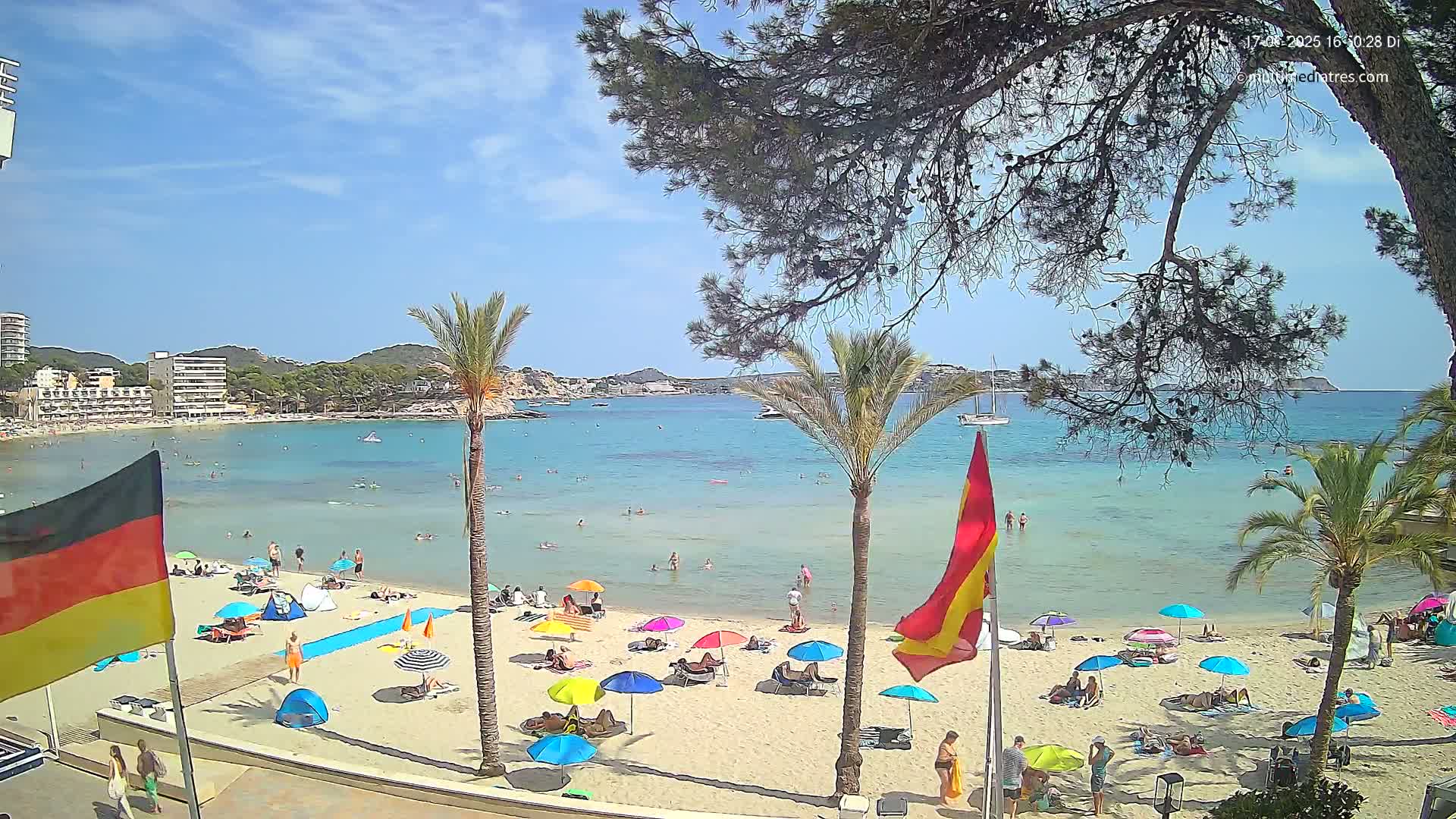 A sunny beach scene shows a calm, clear turquoise ocean with people swimming and sunbathing under colorful umbrellas on a sandy shore, palm trees and buildings are visible in the background.