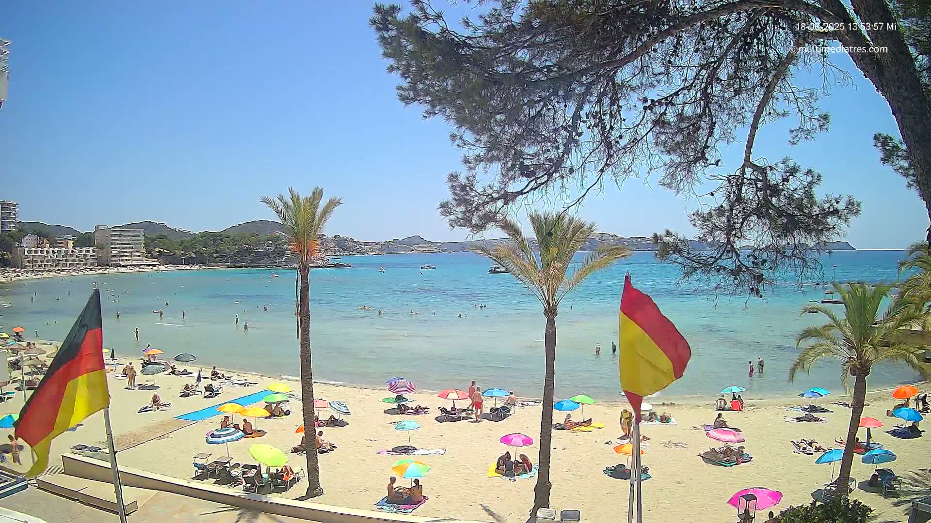 A sunny beach scene shows a sandy shore with colorful umbrellas and people relaxing, calm clear water, and palm trees framing a view of a bay and distant hills.