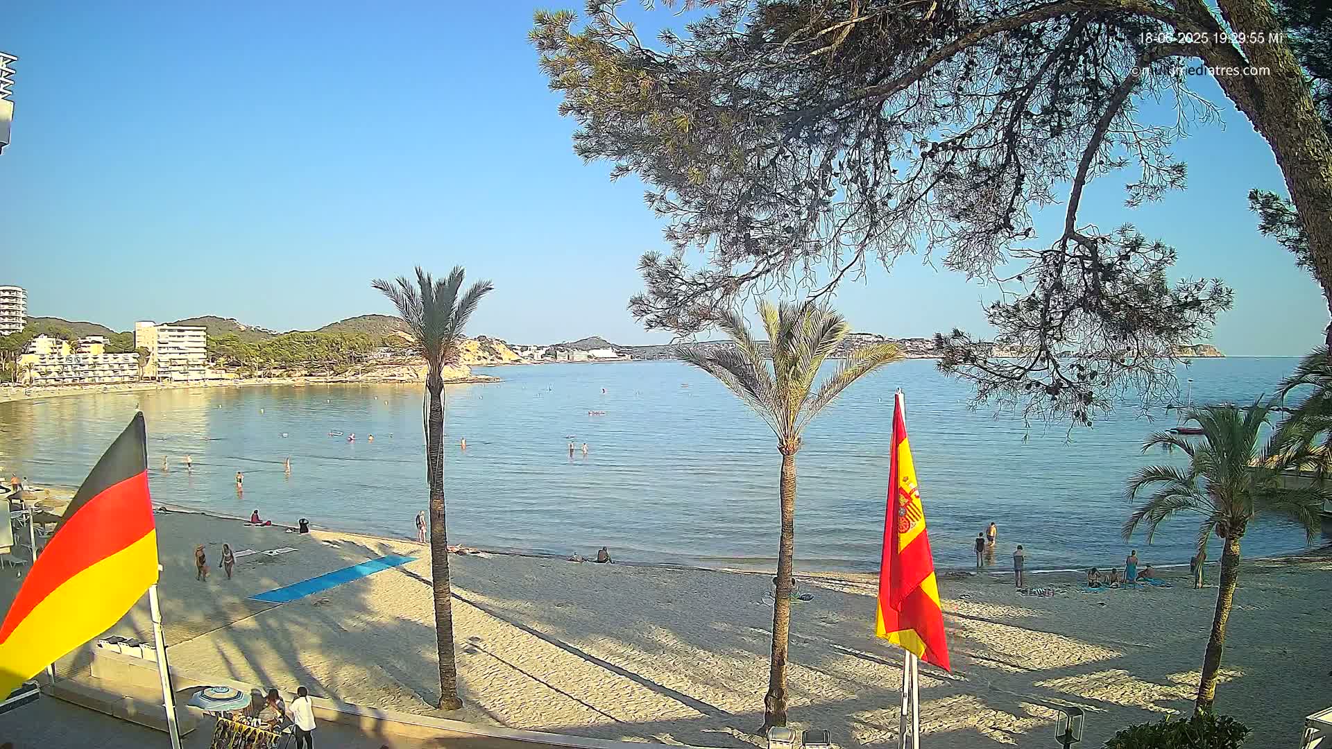 A sandy beach with calm, clear water, palm trees, and buildings in the background under a sunny sky.