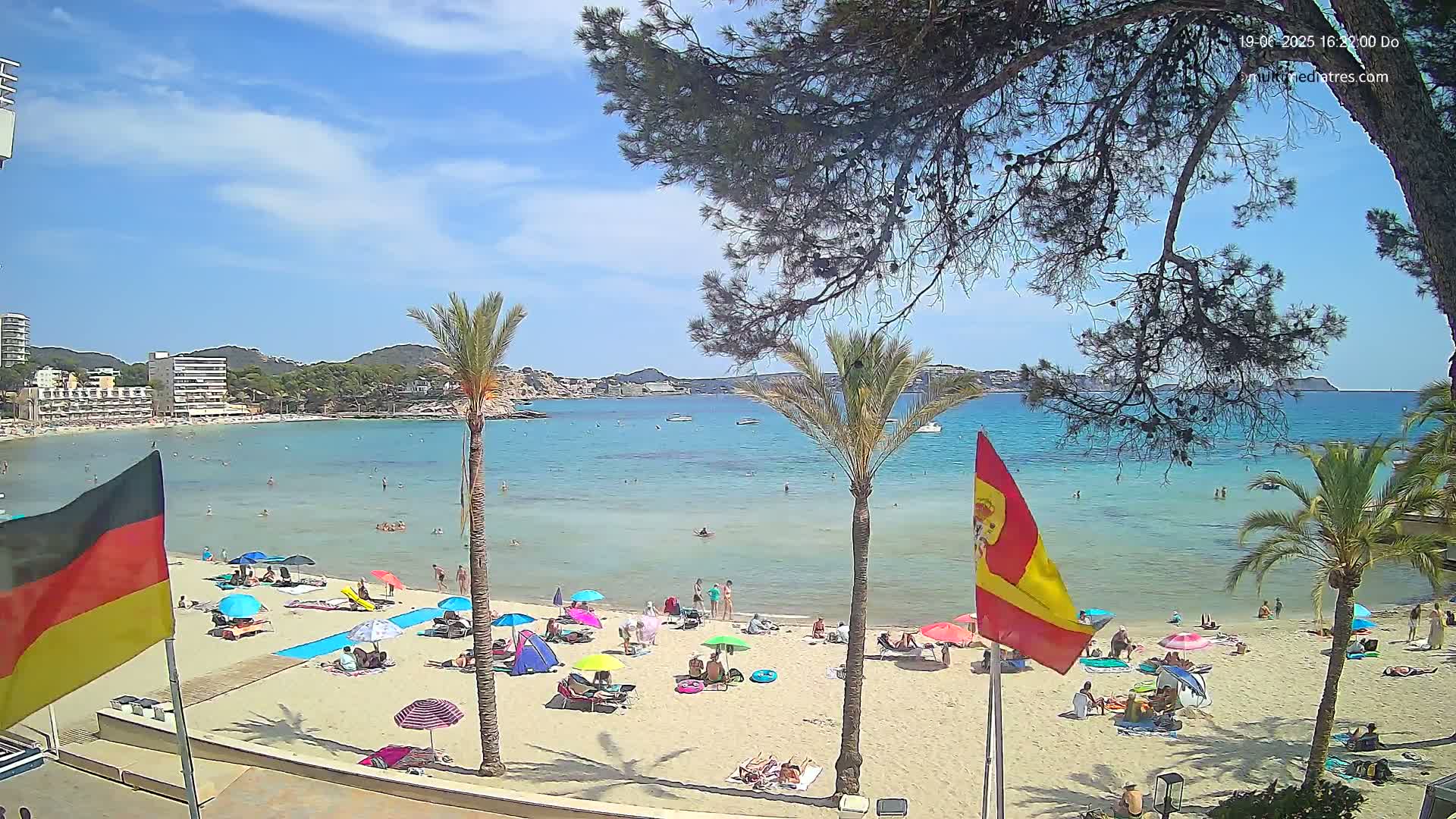 A sunny beach scene shows a calm, clear turquoise ocean, a sandy shore with people relaxing under umbrellas, and palm trees in the foreground, with buildings and hills visible in the distance.