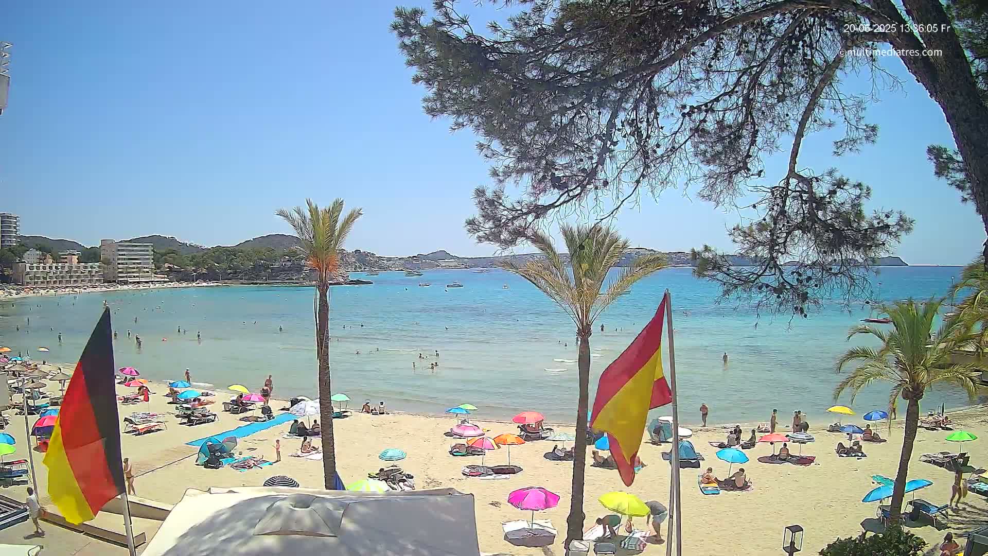 A sunny beach scene shows a calm, clear turquoise ocean with people swimming and sunbathing under colorful umbrellas on a sandy shore, palm trees, and buildings in the background.